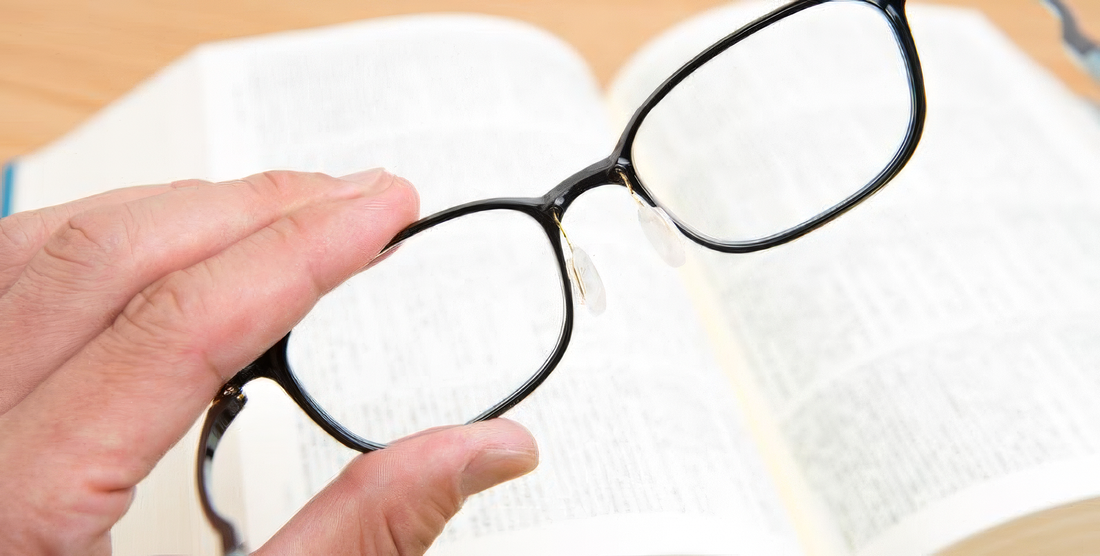 Mano regge un paio di occhiali davanti a un libro aperto per metterne a fuoco il testo