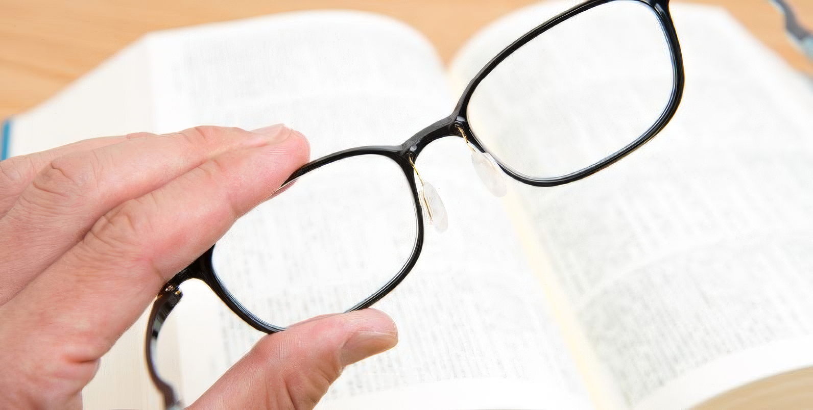 Mano regge un paio di occhiali davanti a un libro aperto per metterne a fuoco il testo