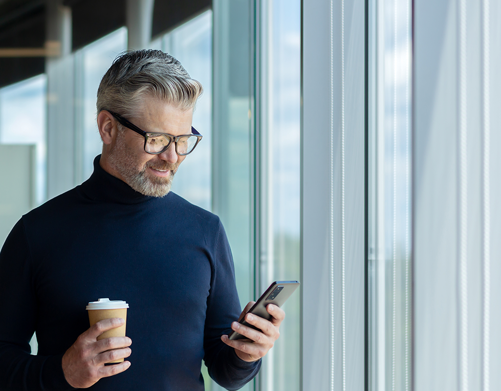 Uomo dai capelli grigi con occhiali consulta lo smartphone tenendo un caffè