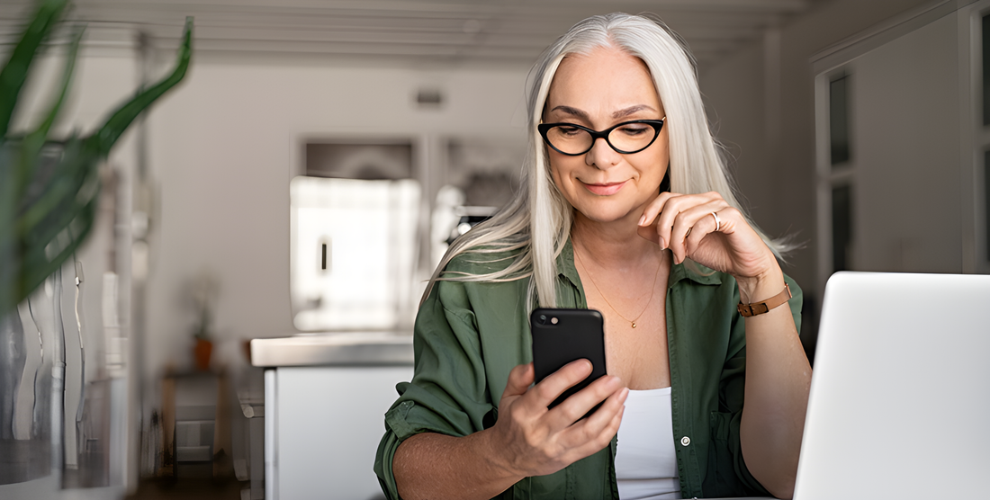 Donna dai capelli grigi con occhiali da vista usa lo smartphone accanto al portatile