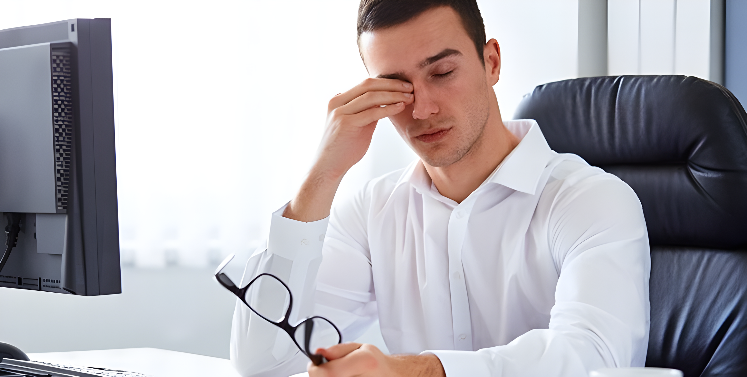Uomo in ufficio si strofina gli occhi affaticati tenendo gli occhiali davanti al monitor