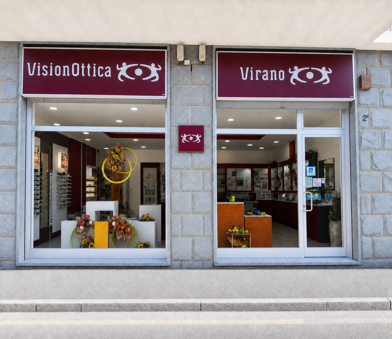 Facciata del negozio VisionOttica Virano di Torino su strada con due vetrine separate, insegne bordeaux con scritta bianca, decorazioni floreali in vetrina e porta a vetri laterale