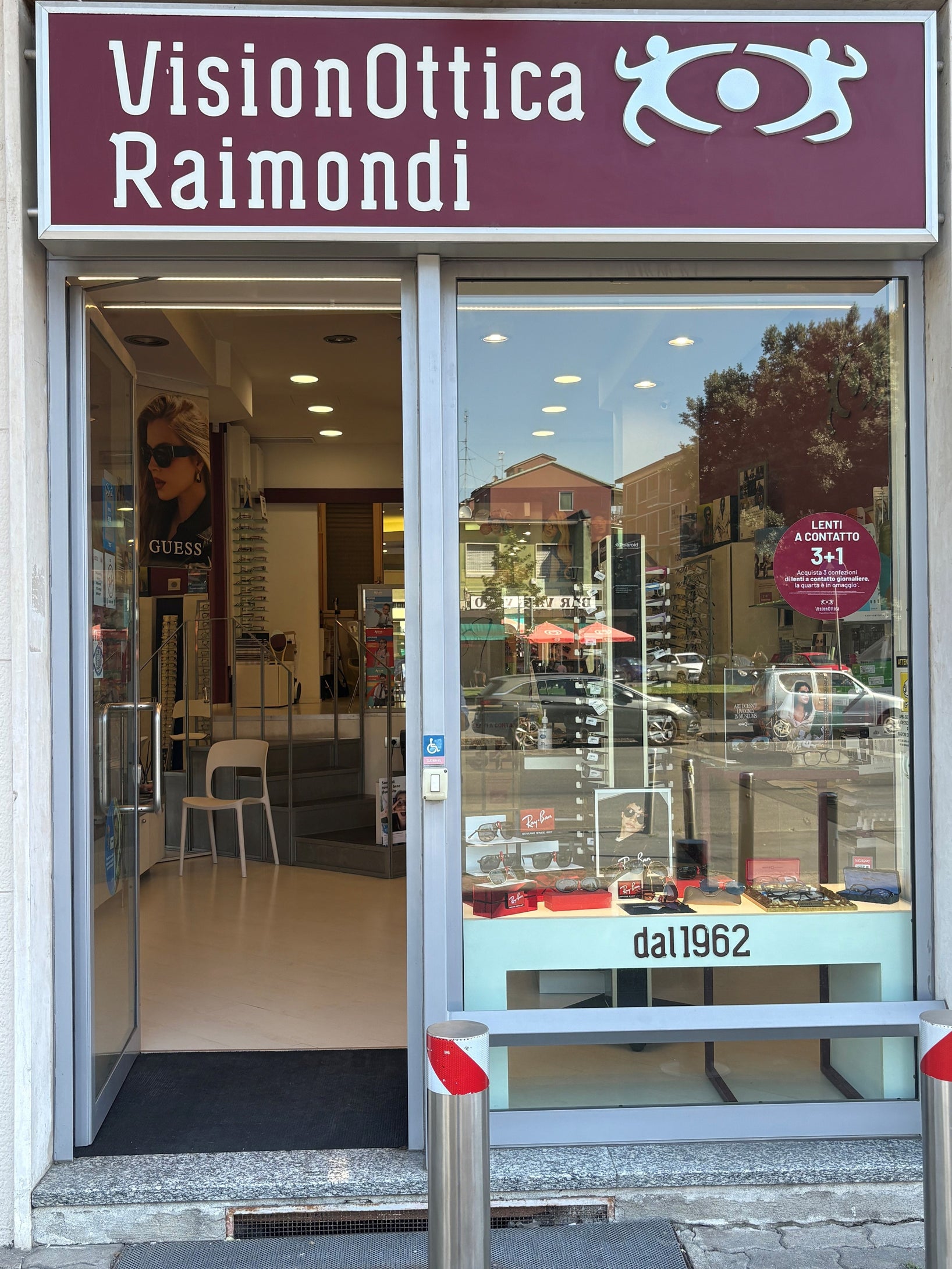 Ingresso del negozio VisionOttica Raimondi di Milano su strada con porta a vetri aperta, insegna bordeaux con scritta bianca, vetrina con occhiali da sole e adesivo 'dal 1962'