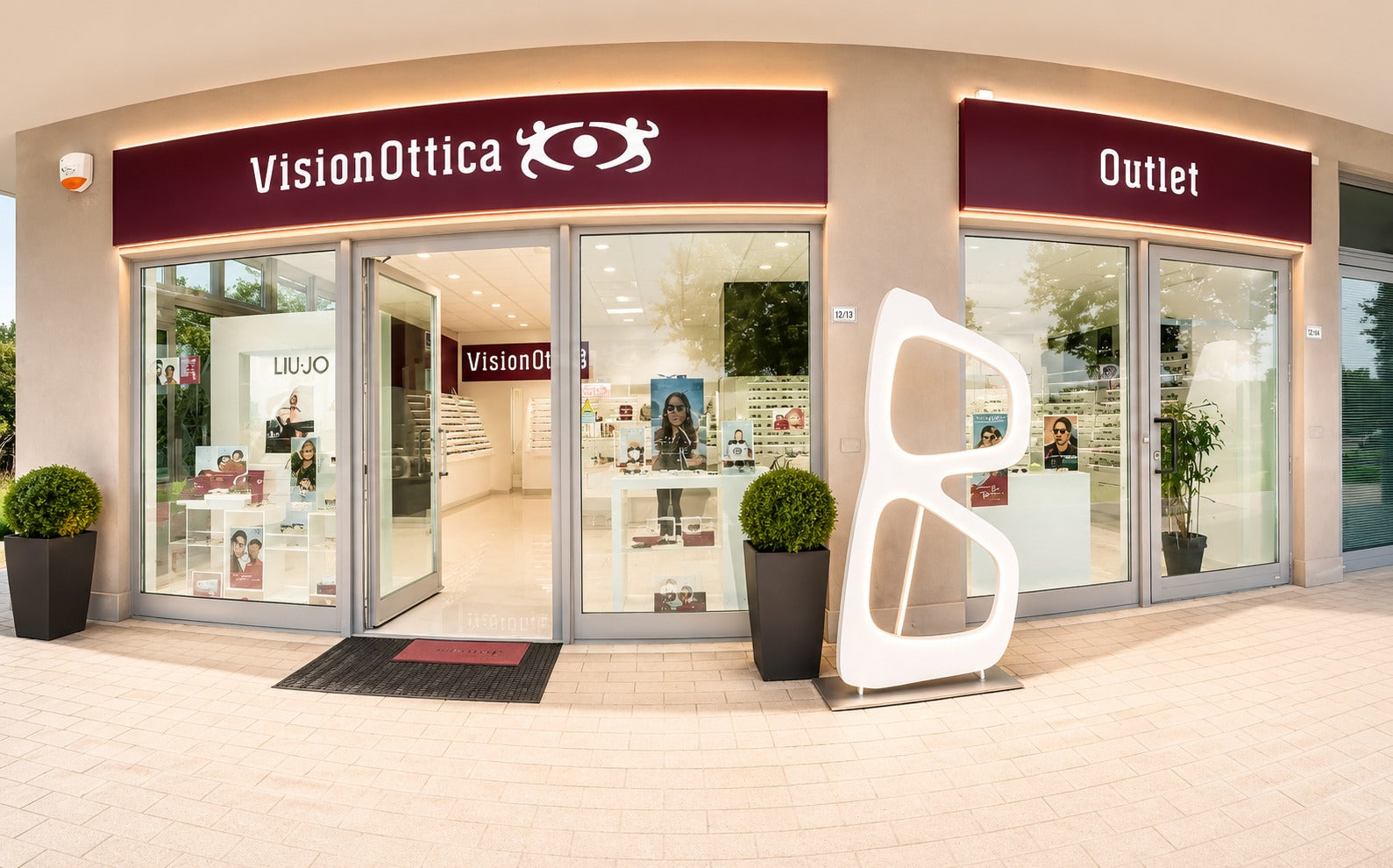 Facciata del negozio VisionOttica Outlet di Castel Guelfo di Bologna in centro commerciale con ampia vetrata a tre moduli, insegne bordeaux e scultura decorativa a forma di occhiale bianco all'esterno