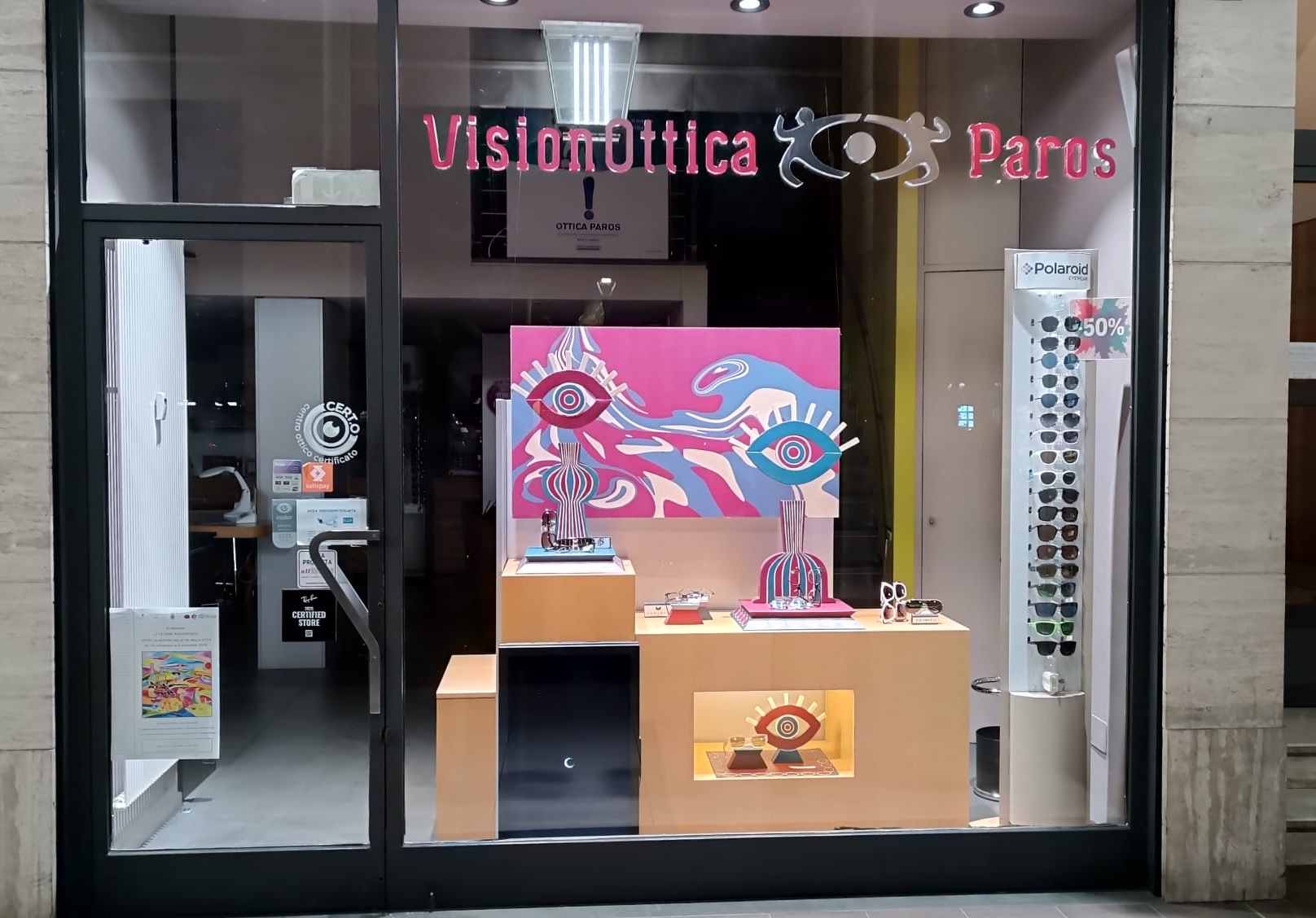 Vetrina del negozio VisionOttica Paros di Canelli in centro storico con allestimento pop-art colorato, espositore Polaroid con occhiali da sole e insegna bordeaux con lettere in rilievo rosa