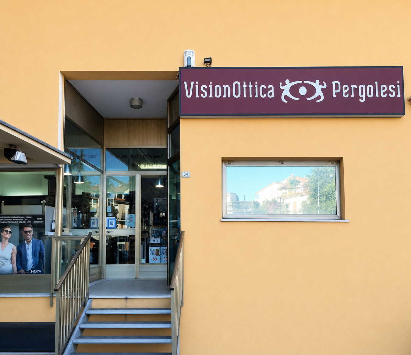 Ingresso del negozio VisionOttica Pergolesi di Osimo su facciata giallo ocra con insegna bordeaux, porta a vetri con gradini di accesso e finestra laterale