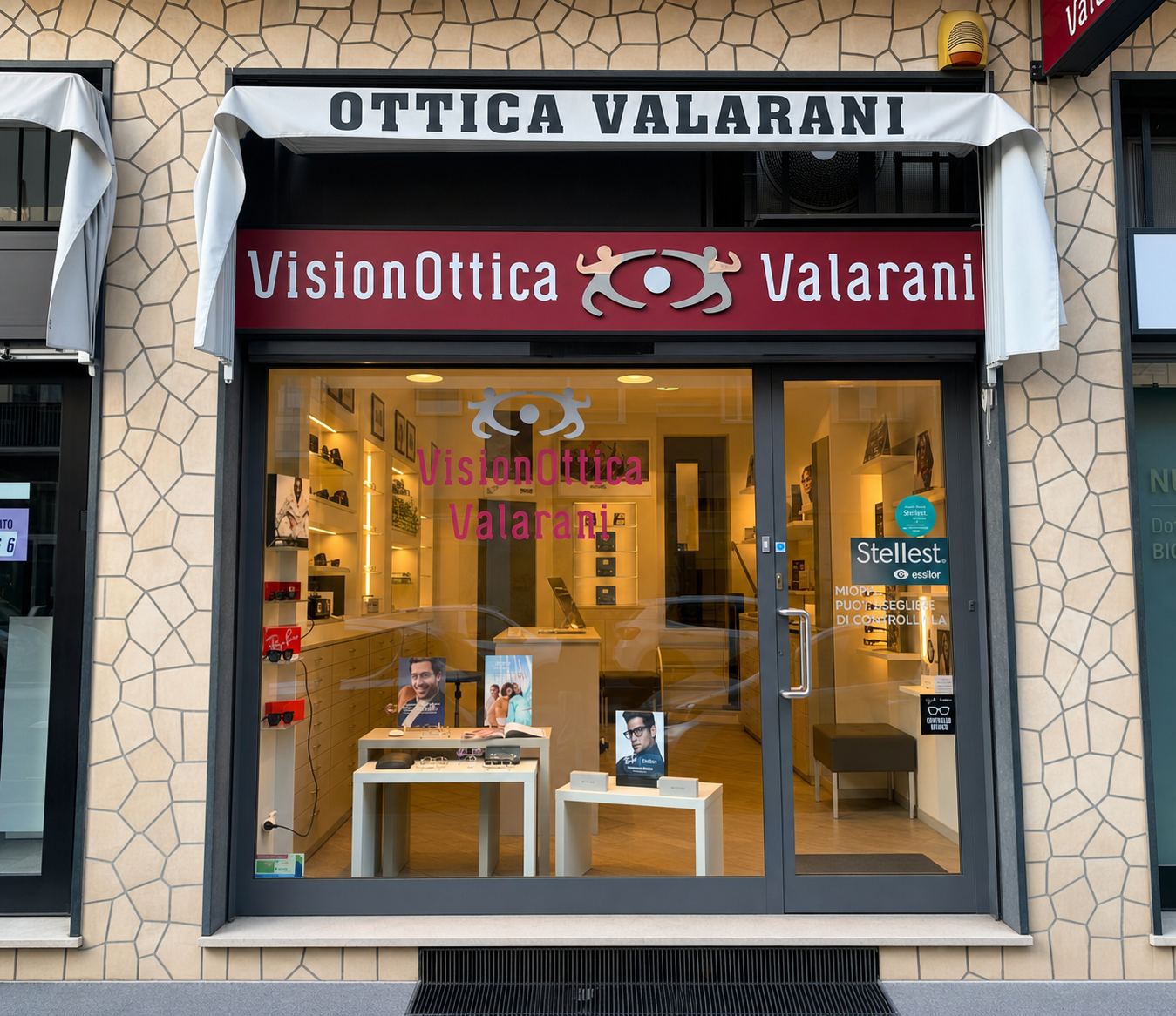 Facciata del negozio VisionOttica Valarani di Milano su strada con vetrina ampia a pavimento, insegna superiore bianca con 'Ottica Valarani' e fascia bordeaux con logo VisionOttica, espositori interni con occhiali e banner Stellest