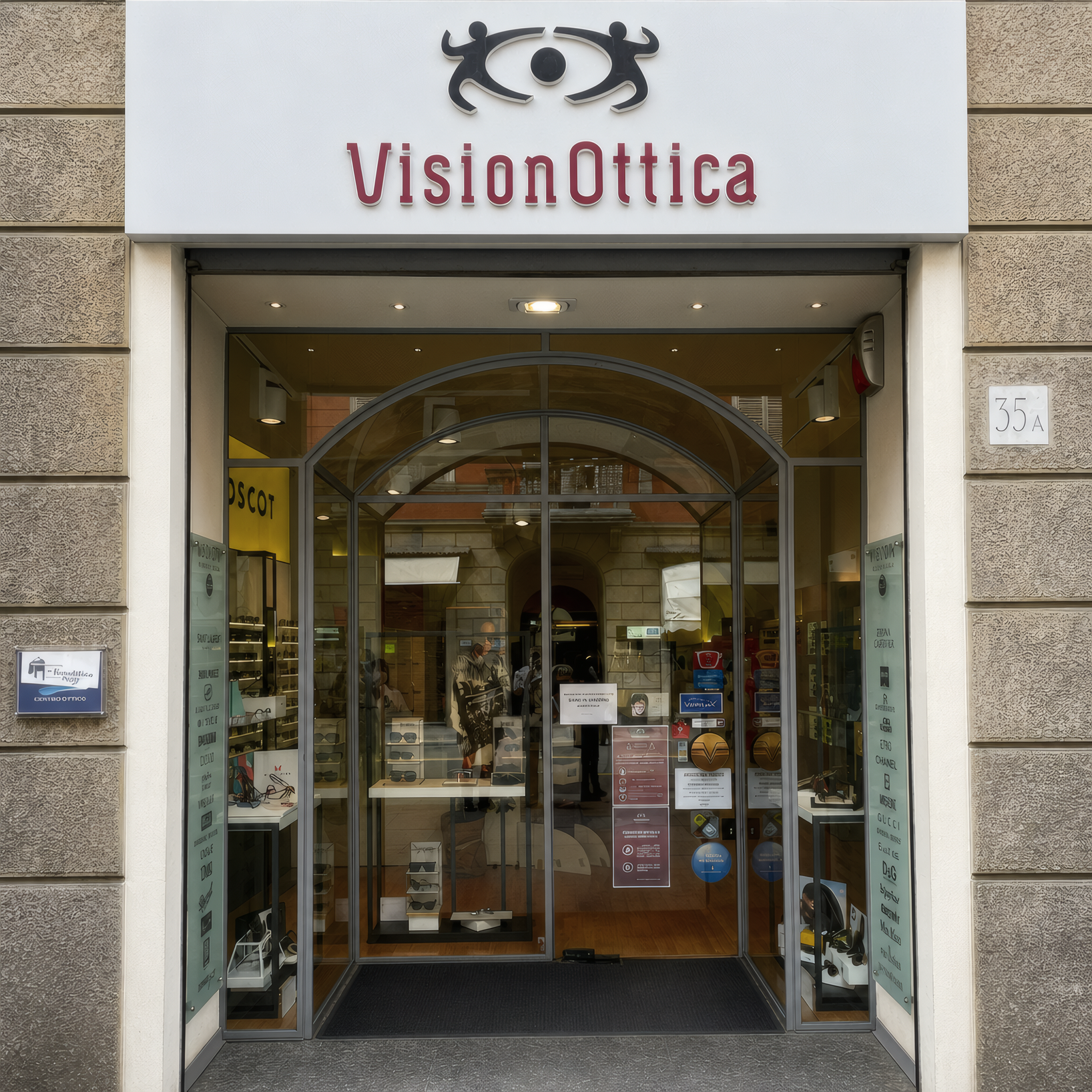 Ingresso del negozio VisionOttica di Parma in centro storico sotto portici con arco in pietra, insegna bordeaux con logo e porta a vetri aperta verso l'interno illuminato