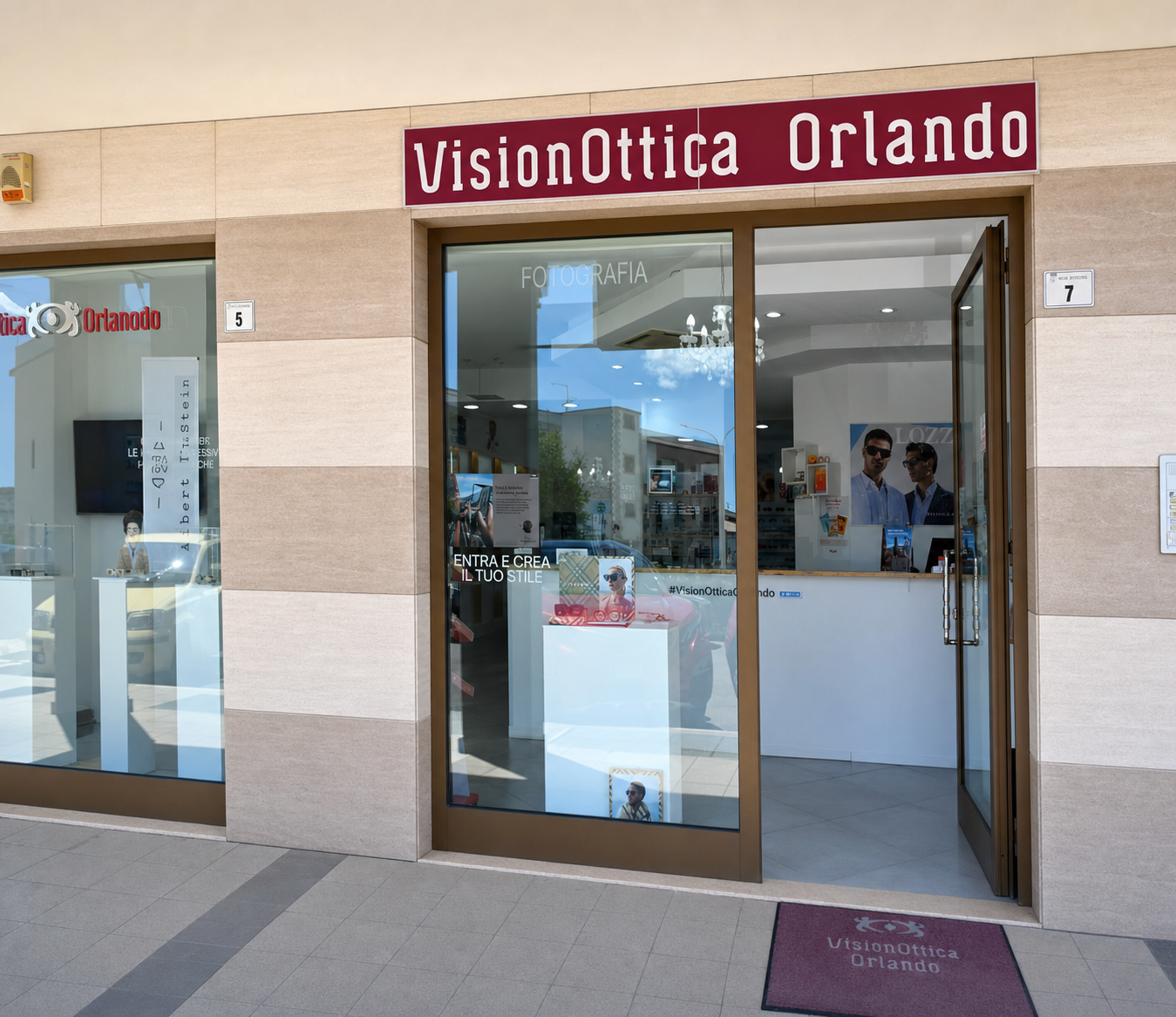 Ingresso del negozio VisionOttica Orlando di Policoro su strada con portici, vetrina laterale con occhiali esposti, porta a vetri e banner 'Entra e fai il tuo stile' sul pavimento all'entrata