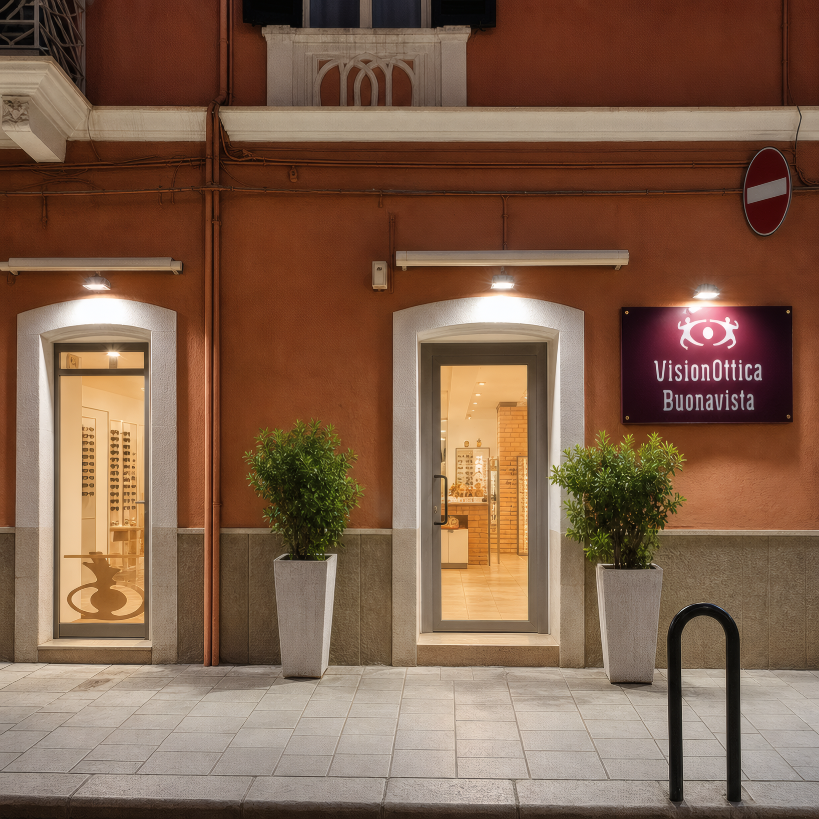 Ingresso del negozio VisionOttica Buonavista di Monopoli in centro storico su edificio color terracotta con porta a vetri aperta, due vasi con alberi decorativi ai lati e insegna bordeaux laterale illuminata di sera