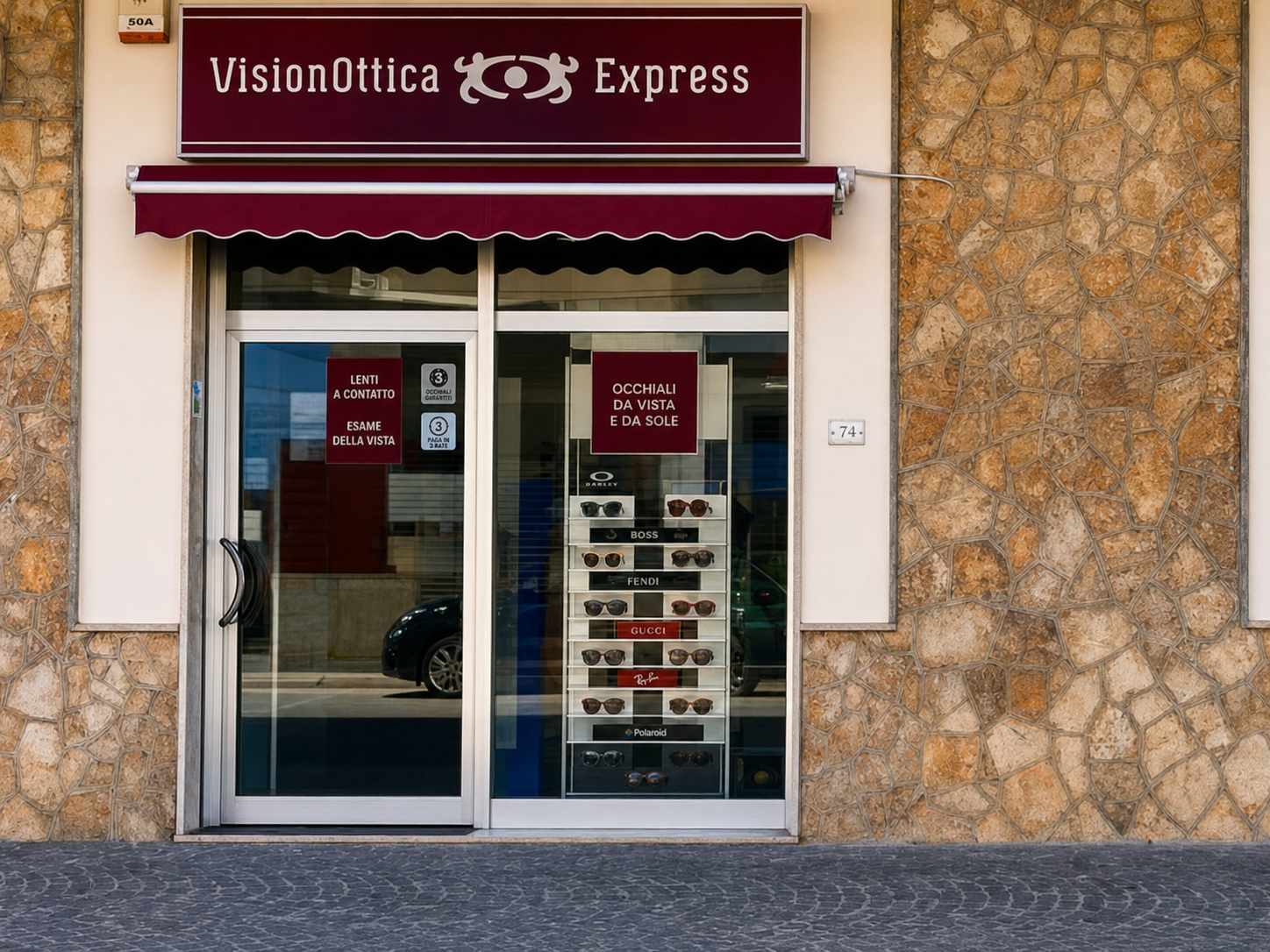 Facciata del negozio VisionOttica Express di Mondragone su strada con muro in pietra, tenda bordeaux a strisce, porta a vetri con adesivi promozionali e vetrina con espositore occhiali da sole di brand