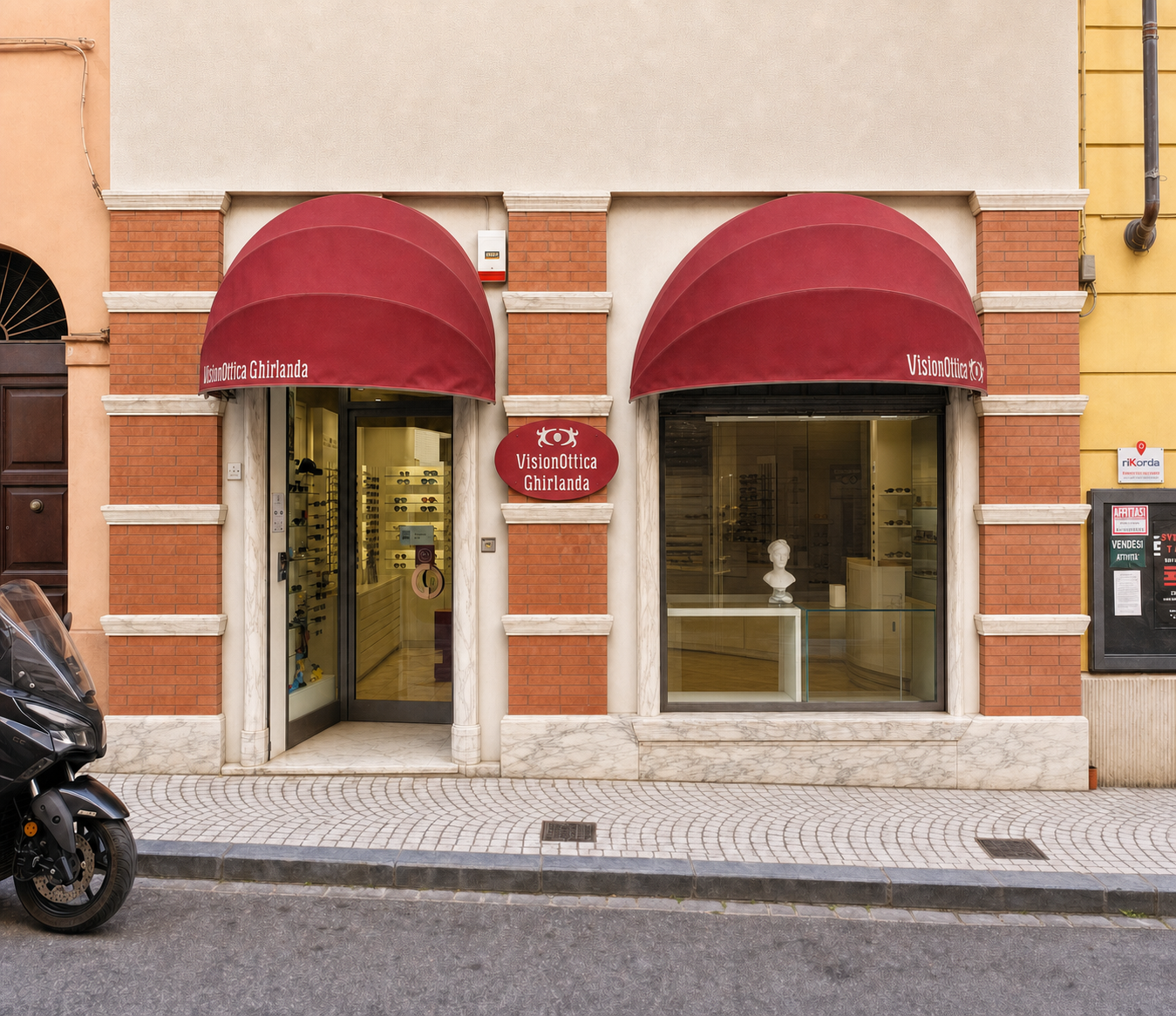 Facciata del negozio VisionOttica Ghirlanda di Carrara in centro storico su palazzo in mattoni rossi con due tende a cupola bordeaux, due vetrine con espositori di montature e insegna ovale centrale