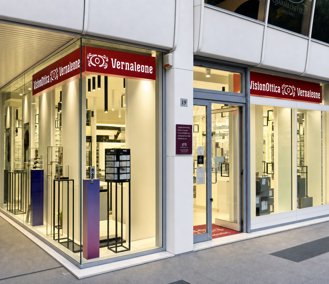 Facciata del negozio VisionOttica Vernaleone di Lecce in centro commerciale moderno con ampia vetrata su angolo, insegne bordeaux su due lati, espositori verticali viola con montature all'interno e tappeto rosso all'ingresso