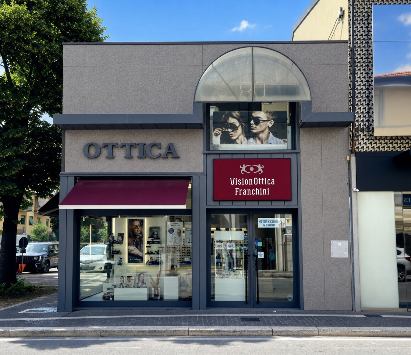 Facciata del negozio VisionOttica Franchini di Arezzo su strada con edificio bimodulare, insegna 'Ottica' in lettere scure a sinistra, tenda bordeaux retratta, insegna bordeaux VisionOttica Franchini a destra e lightbox con coppia in occhiali