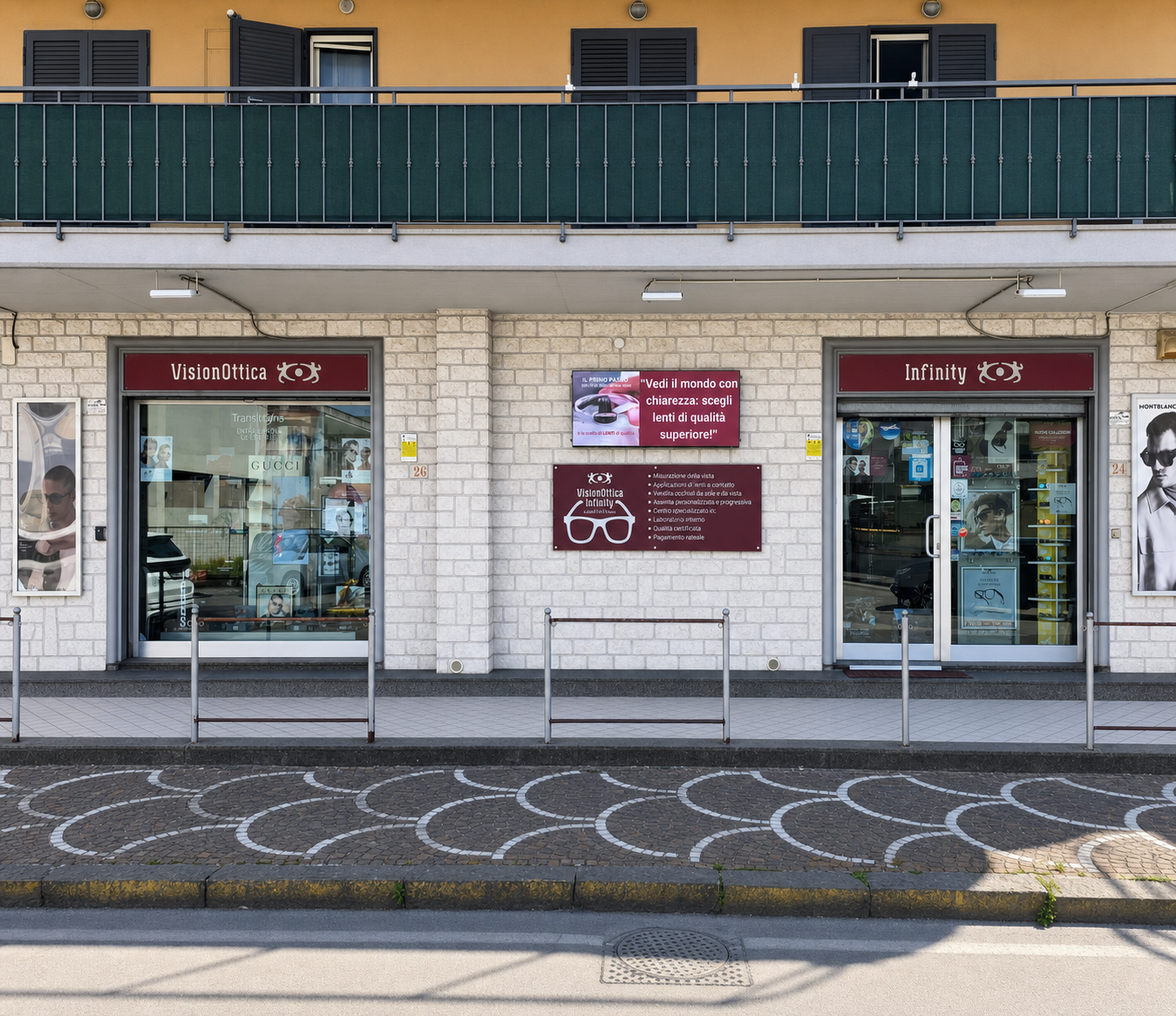 Facciata del negozio VisionOttica Infinity di Scafati su strada con edificio a due moduli, insegne bordeaux separate, pannello promozionale centrale con slogan e icona occhiale, vetrine con espositori e poster laterali