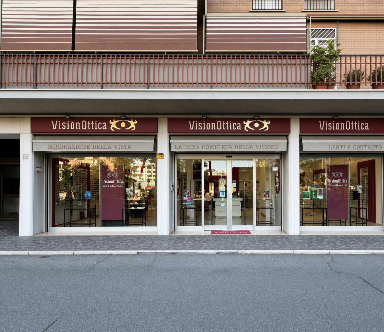 Facciata del negozio VisionOttica di Roma su strada con tre moduli affiancati, insegne bordeaux con slogan 'Misurazione della Vista', 'La cura completa della visione' e 'Lenti a contatto', porta centrale a vetri e due vetrine laterali con montature