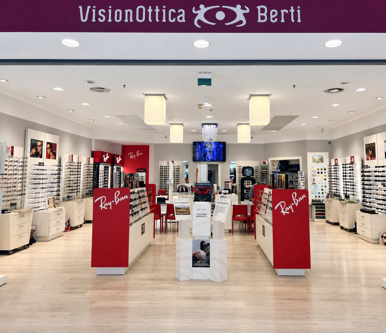 Interno del negozio VisionOttica Berti di Trebaseleghe in centro commerciale con ampio showroom, espositori Ray-Ban centrali in rosso, scaffalature perimetrali con montature, insegna bordeaux e illuminazione a soffitto