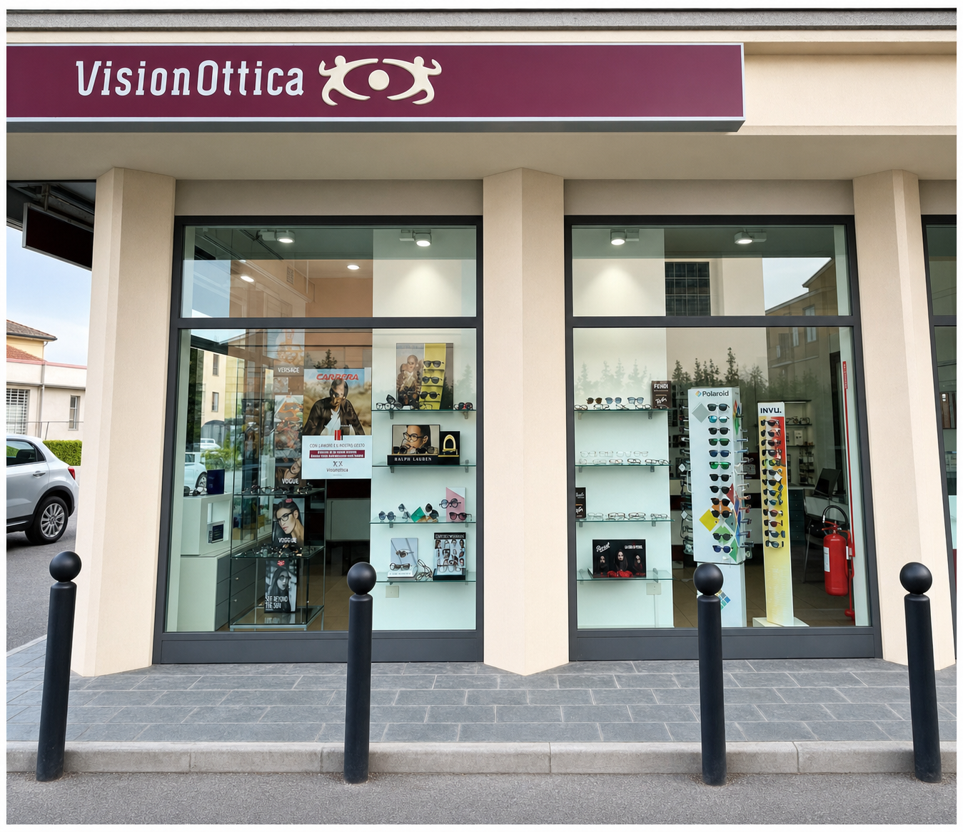 Vetrina del negozio VisionOttica di Colorno su strada con grande vetrata a due moduli, insegna bordeaux, espositori con occhiali da sole e montature visibili attraverso il vetro e bollard neri sul marciapiede