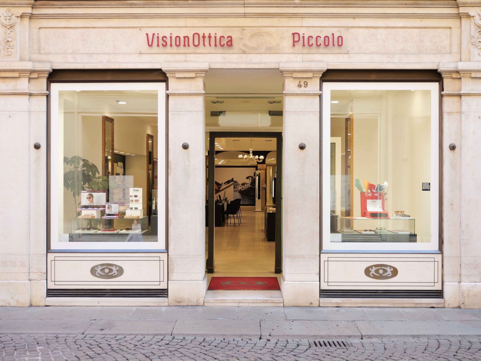 Ingresso del negozio VisionOttica Piccolo di Vicenza in centro storico su palazzo classico in pietra chiara con porta centrale aperta, due vetrine laterali ad arco con occhiali esposti e insegna rossa con lettere in rilievo