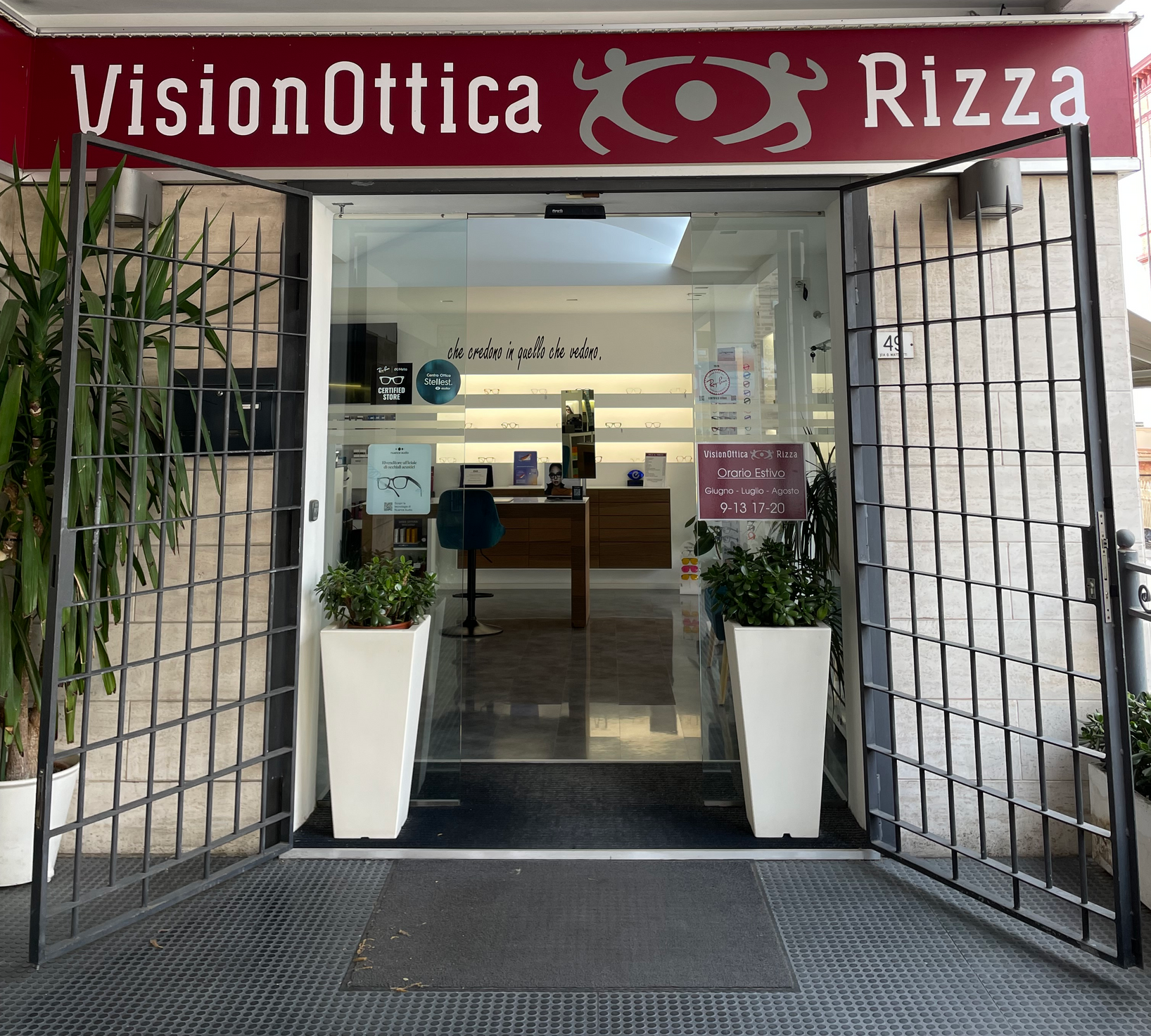 Ingresso del negozio VisionOttica Rizza di Castelfidardo su strada con cancellata metallica aperta, porta a vetri, insegna bordeaux, due vasi con piante all'esterno e bancone interno visibile