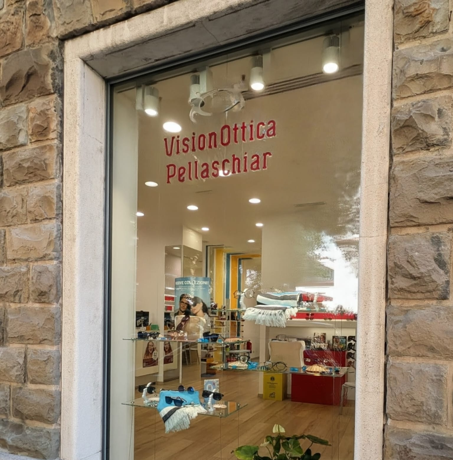 Vetrina del negozio VisionOttica Pellaschiar di Trieste in centro storico incorniciata da muro in pietra a vista, con insegna rossa all'interno, espositori di occhiali e accessori colorati su più ripiani