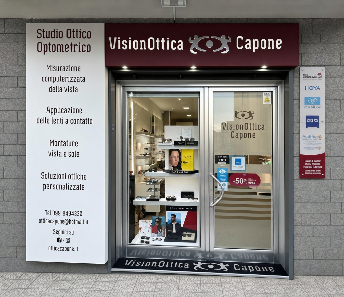 Facciata del negozio VisionOttica Capone di Mottola su strada con insegna bordeaux, pannello laterale bianco con servizi elencati e porta a vetri con vetrina che mostra occhiali, banner promozionale -50% e brand HOYA e ZEISS