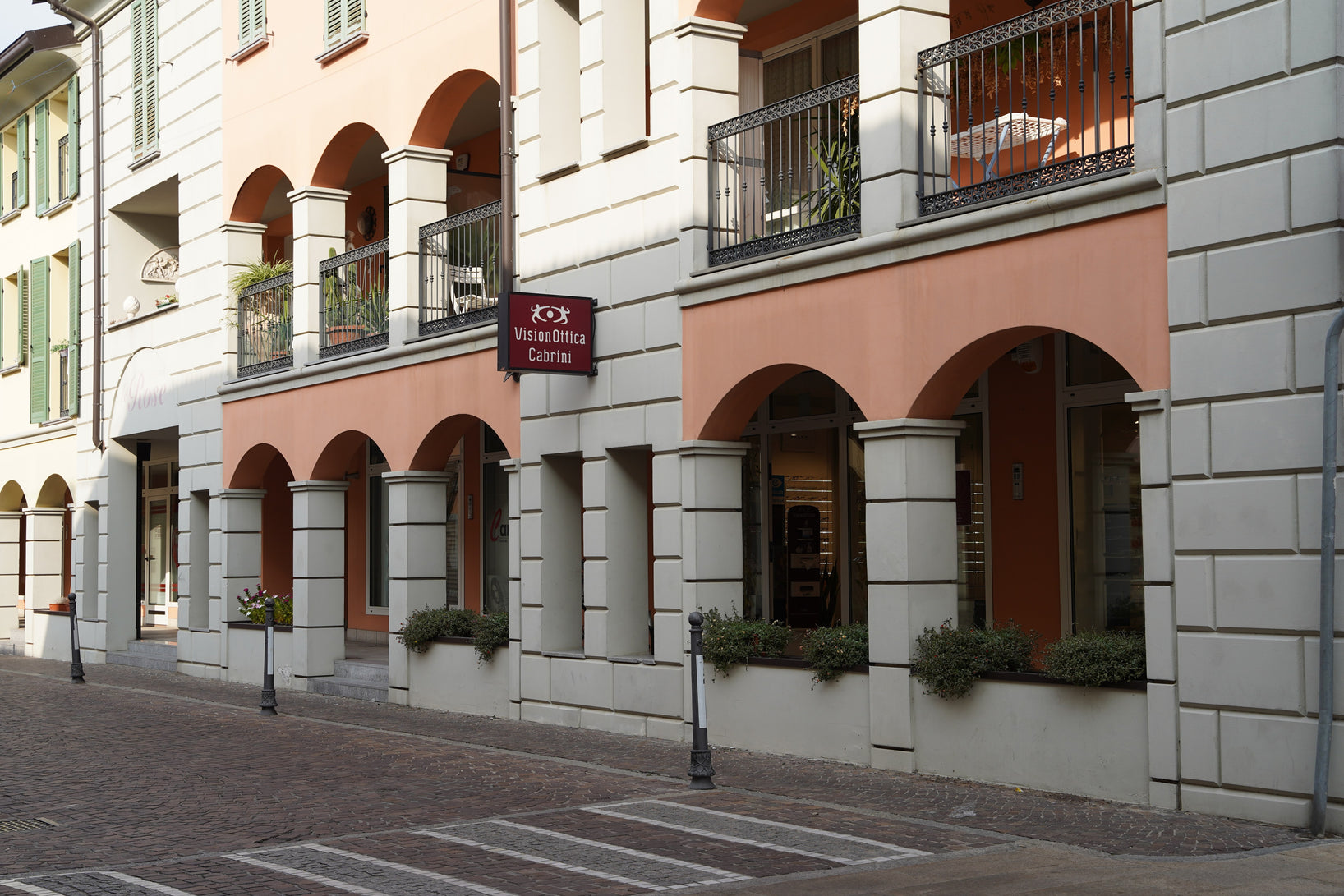 Facciata del negozio VisionOttica Cabrini di Offanengo in centro storico sotto portici con archi in pietra, insegna bordeaux appesa verticalmente, facciata dell'edificio storico color salmone e ingresso ad arco sulla destra