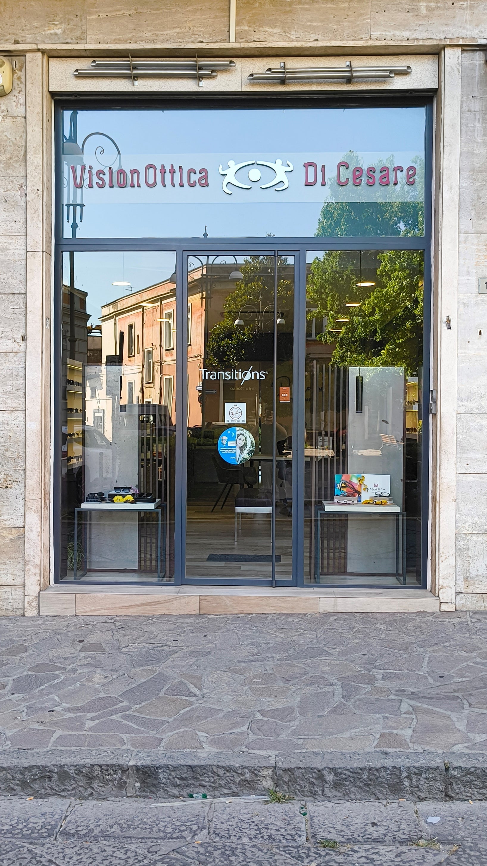 Ingresso del negozio VisionOttica Di Cesare di Maddaloni in centro storico con porta a vetri scorrevole, insegna con logo e adesivo Transitions sulla porta