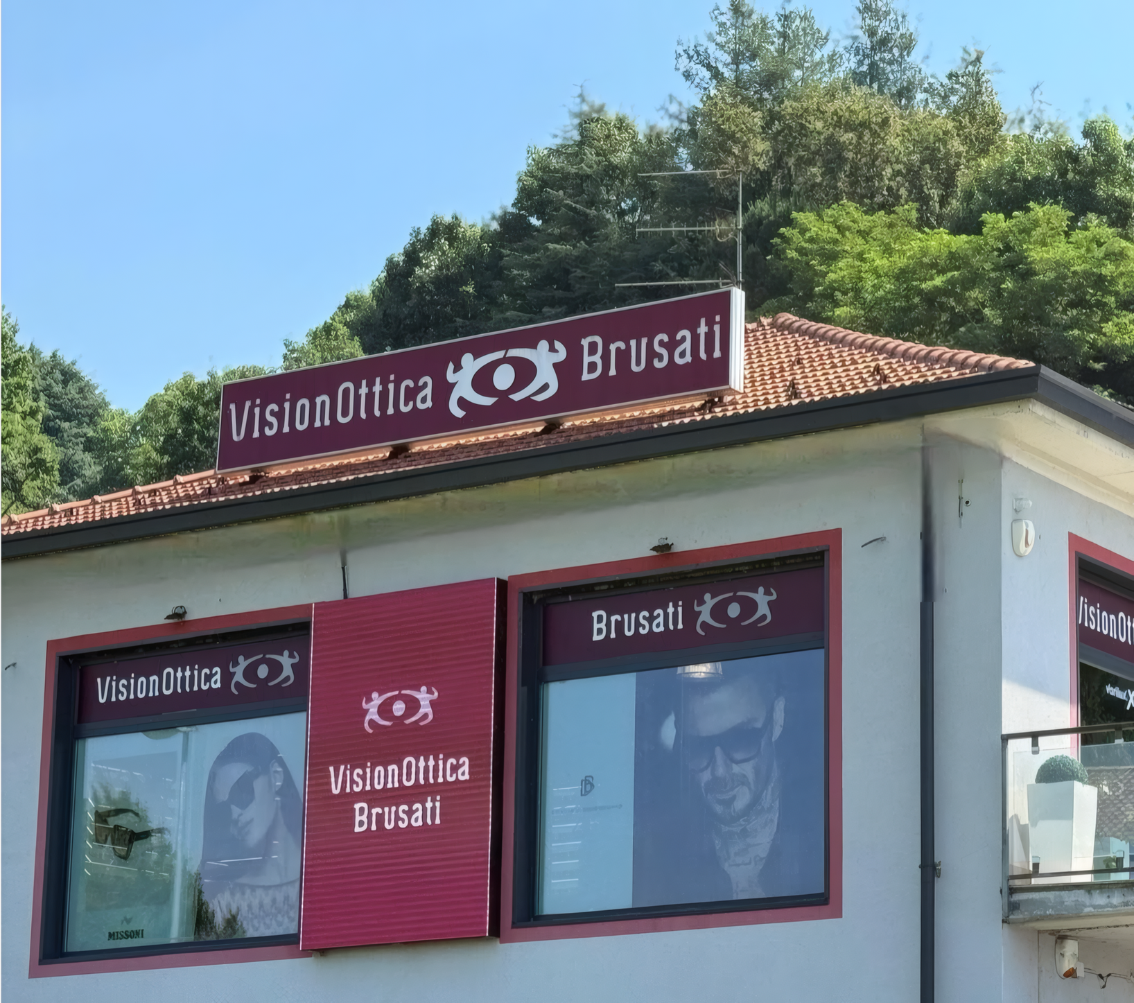 Esterno del negozio VisionOttica Brusati di Solbiate Arno con insegna bordeaux sul tetto, vetrine con immagini promozionali e banner fotografici del brand