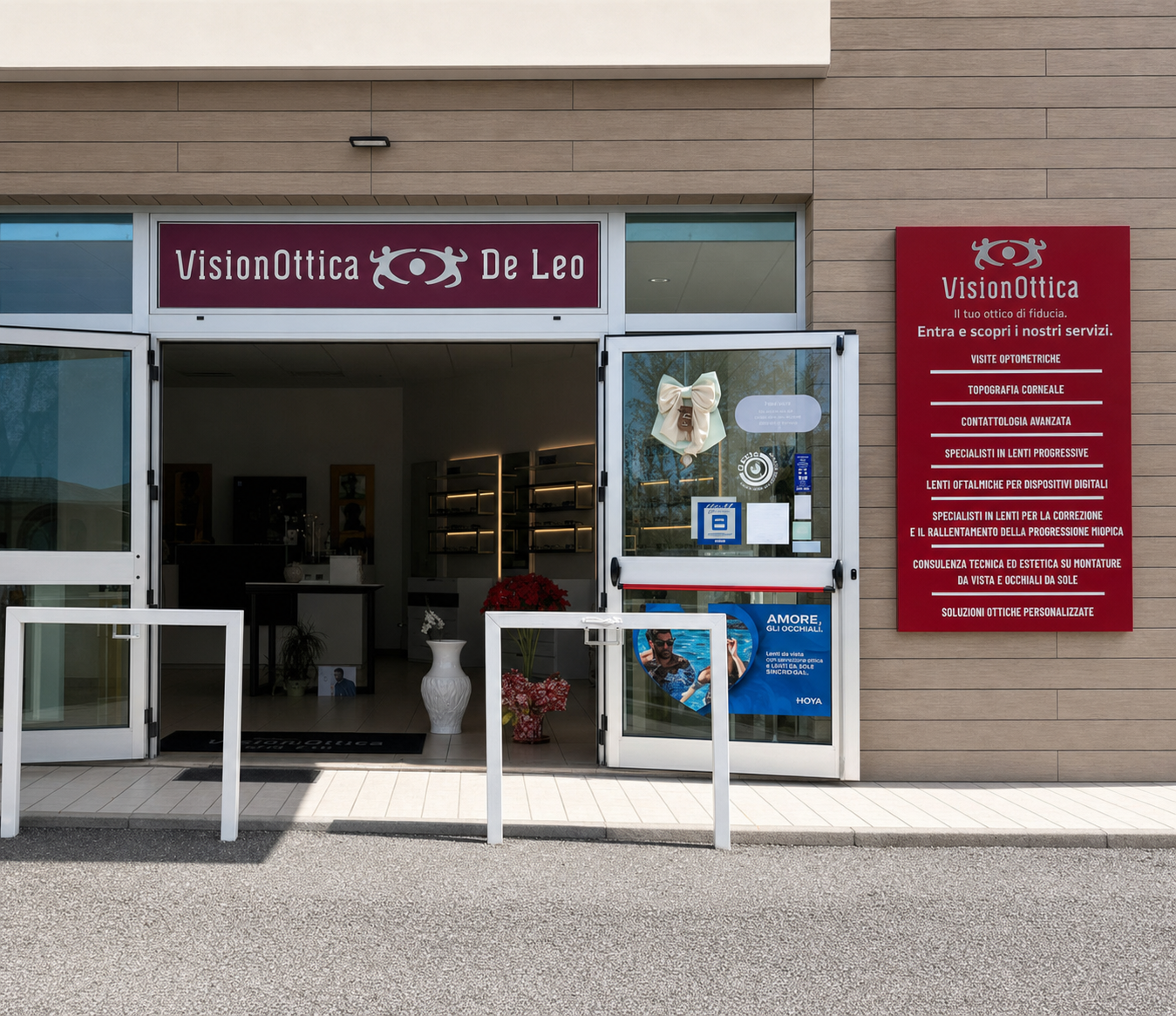 Ingresso del negozio VisionOttica De Leo di Lauria in edificio commerciale moderno con porta a doppio battente aperta, insegna bordeaux e pannello servizi laterale