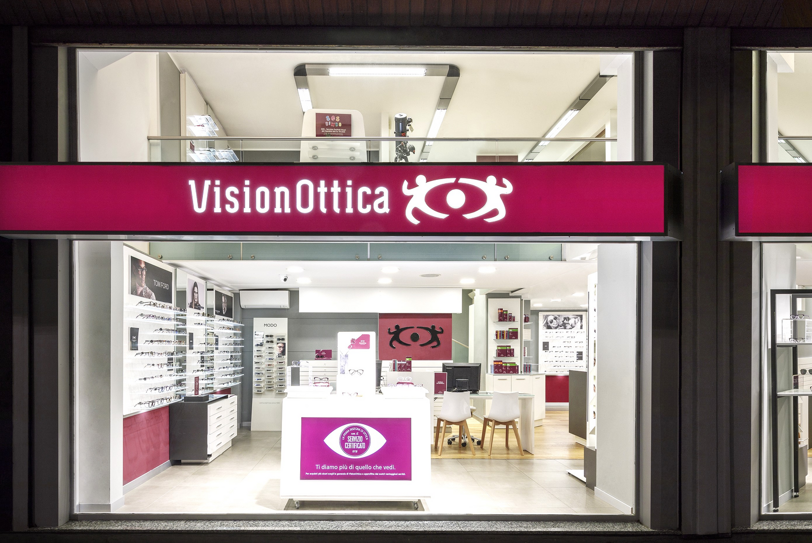 Vetrina del negozio VisionOttica De Angeli di Milano in centro commerciale con insegna fucsia illuminata, bancone centrale bianco, espositori di occhiali su pareti e banner promozionale
