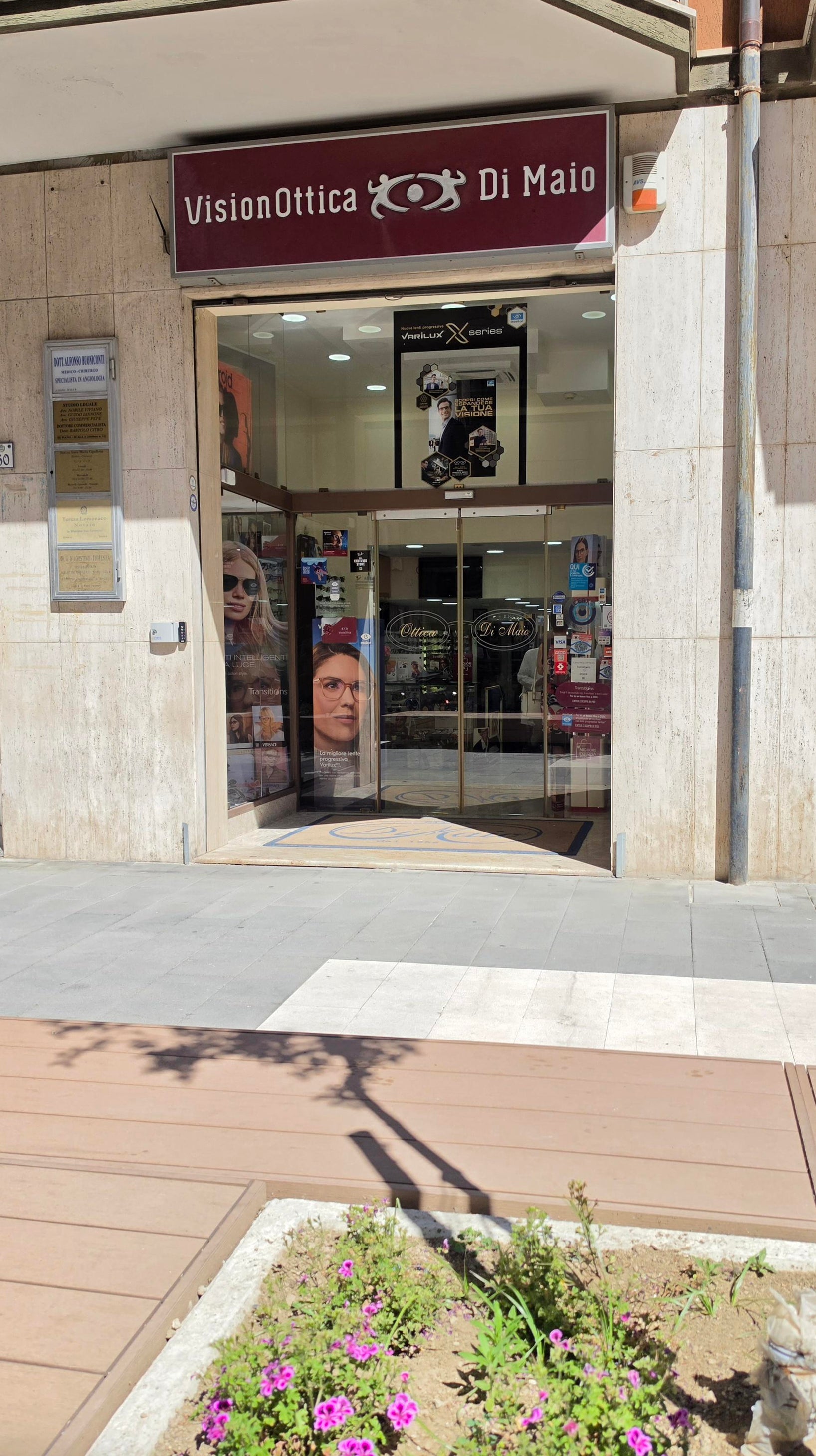 Ingresso del negozio VisionOttica Di Maio di Mercato San Severino su via pedonale con porta automatica a vetri, insegna bordeaux e vetrofanie con immagini di modelle con occhiali