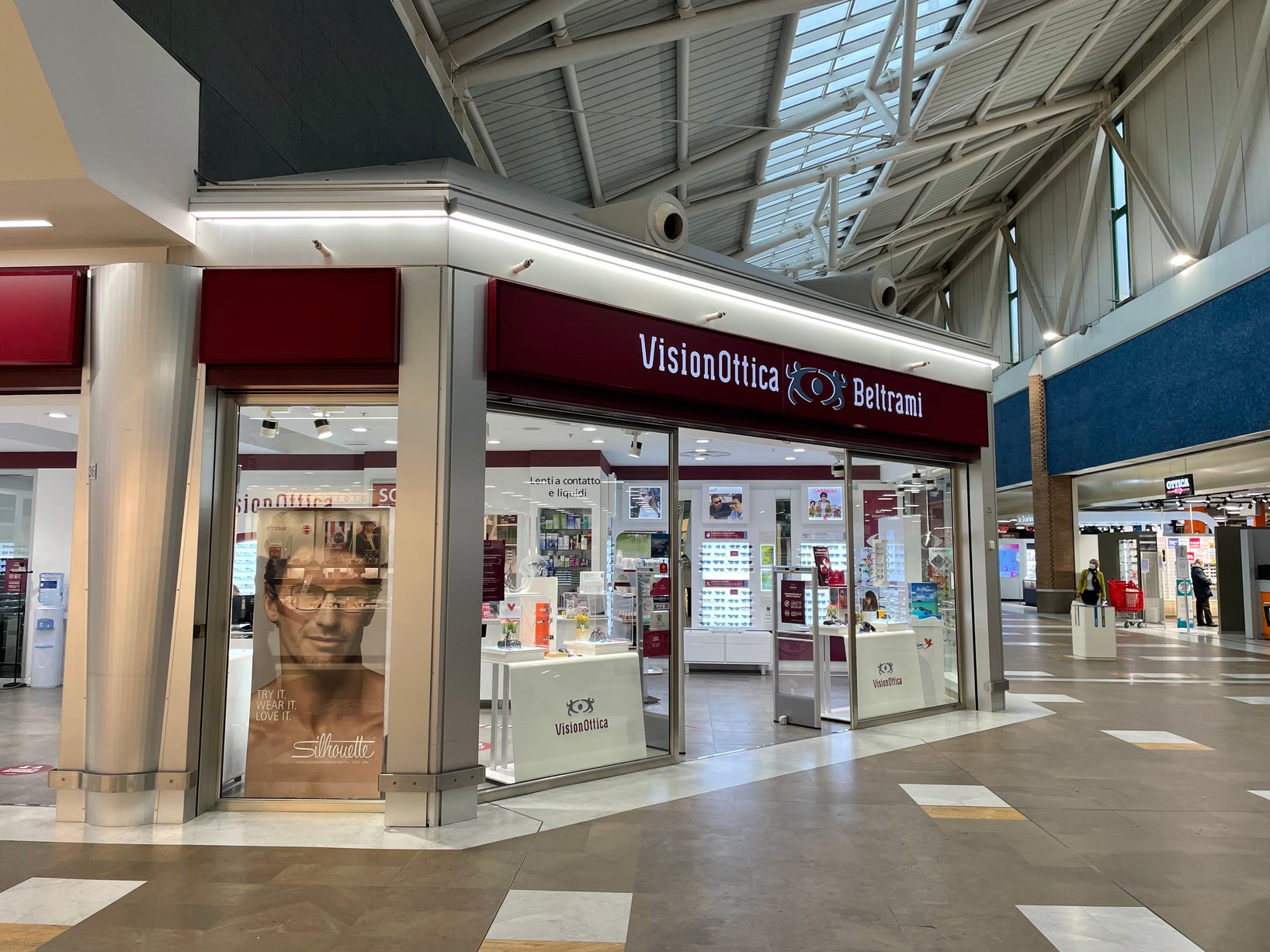 Ingresso del negozio VisionOttica Beltrami di Ferrara in centro commerciale con insegna bordeaux, vetrina con vetrofania fotografica e ampio spazio interno con espositori