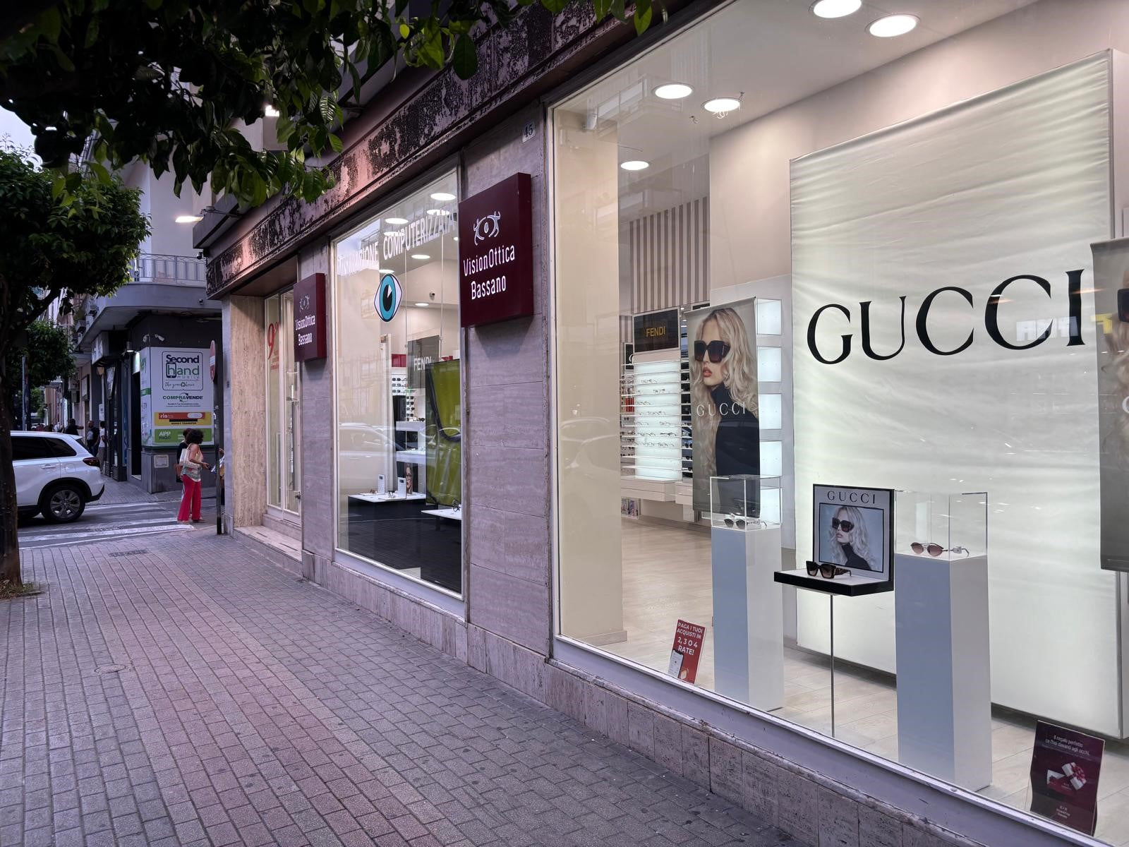 Vetrina del negozio VisionOttica Bassano di Battipaglia su via pedonale alberata con insegna bordeaux, grande display Gucci con occhiali da sole esposti su piedistalli bianchi