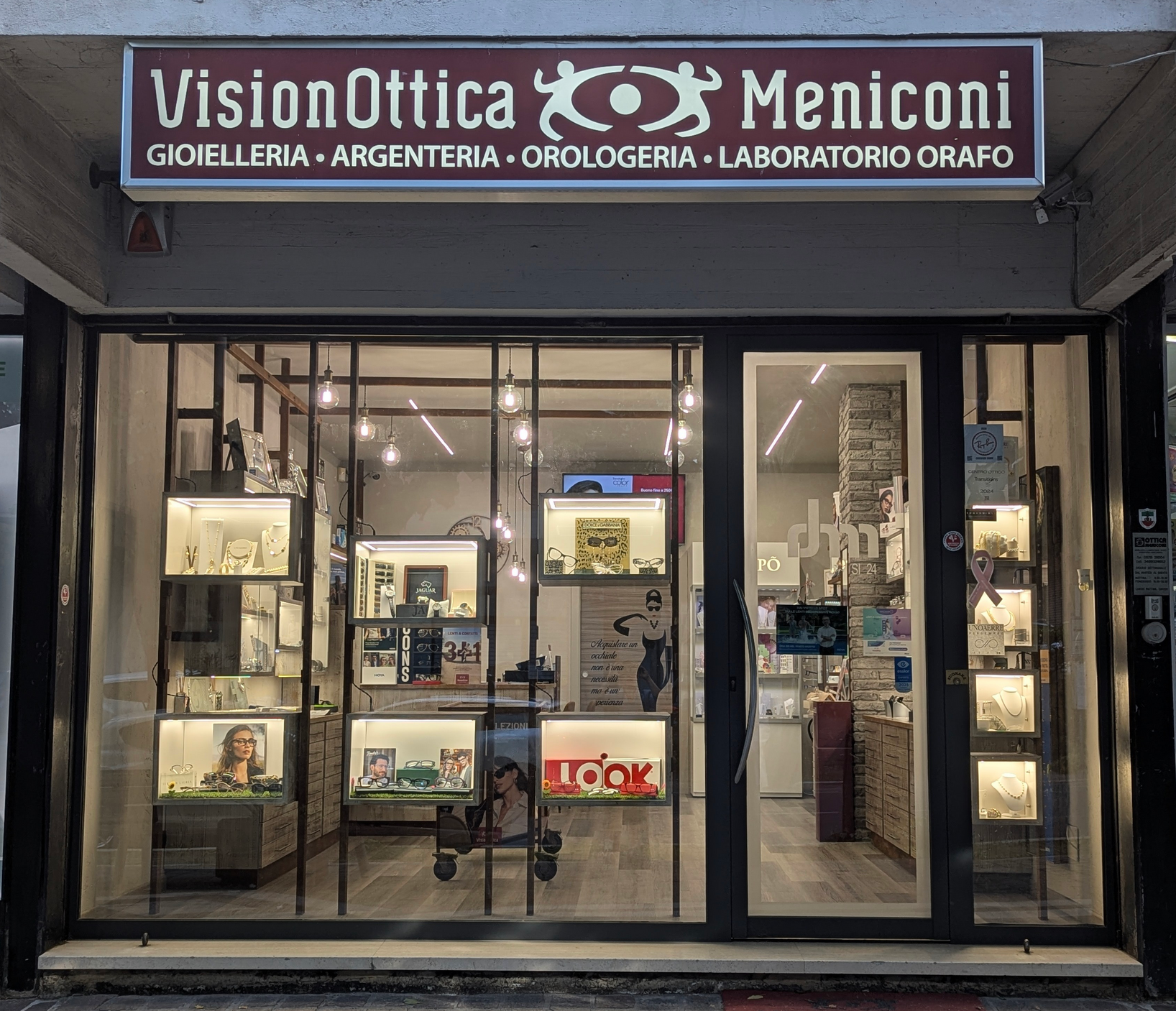 Vetrina notturna del negozio VisionOttica Meniconi di Chianciano Terme su strada con insegna luminosa bianca, espositori illuminati di occhiali e gioielli e pannello Gioielleria Argenteria Orologeria