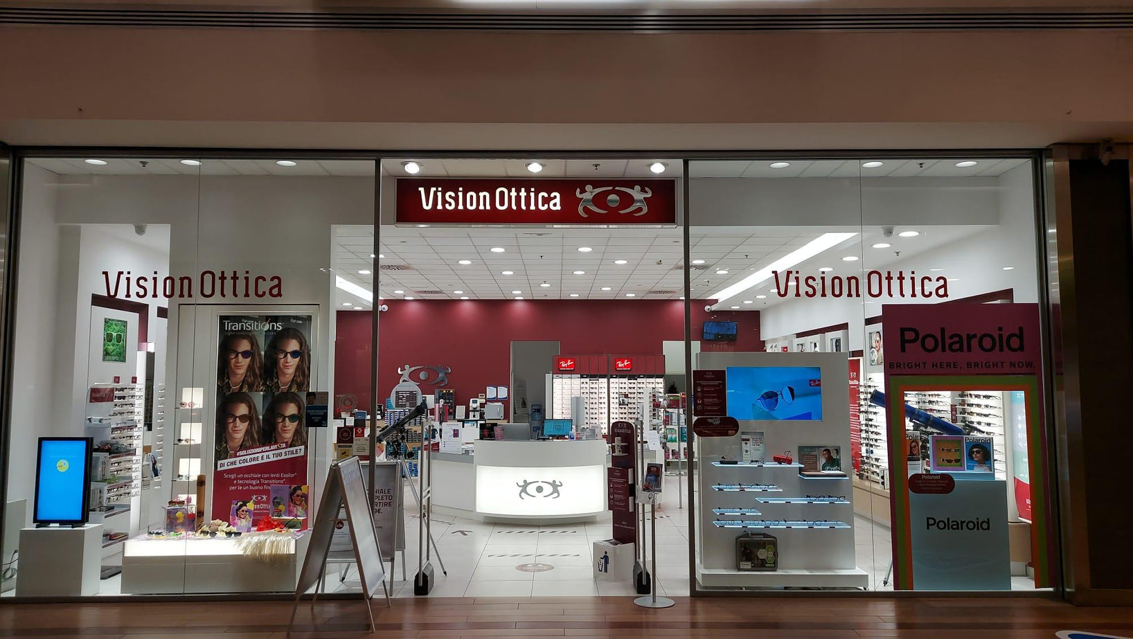 Vetrina del negozio VisionOttica Rho Center di Rho in centro commerciale con insegna rossa, bancone centrale illuminato, espositori di occhiali su pareti e display Polaroid e Transitions