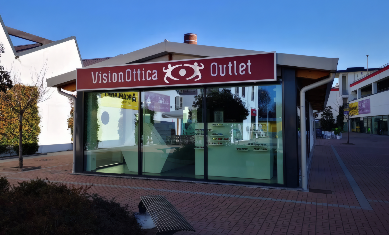 Esterno del negozio VisionOttica Outlet di San Vittore Olona in area commerciale con edificio autonomo a vetrate, insegna bordeaux con scritta Outlet e panchina esterna