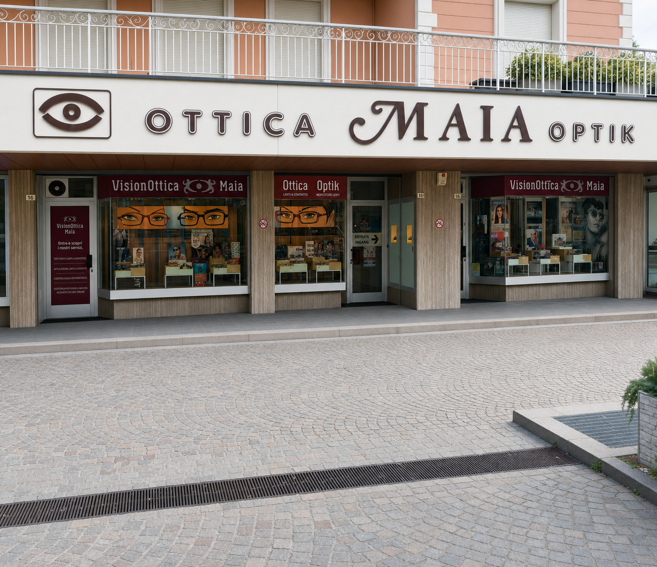 Facciata del negozio VisionOttica Maia di Merano su piazza lastricata con insegna principale Ottica Maia Optik, tre vetrine con espositori di occhiali e insegne bordeaux VisionOttica