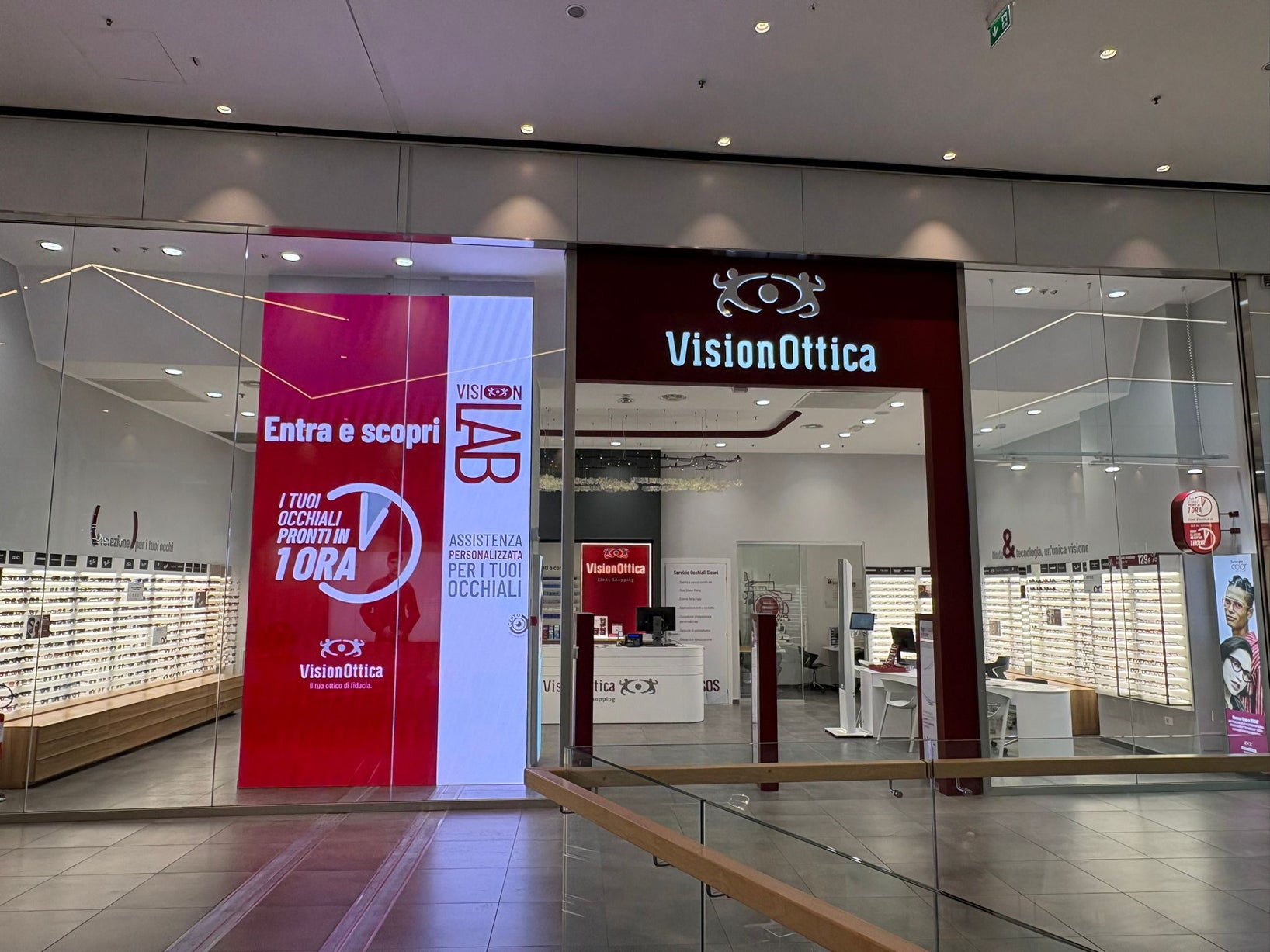 Ingresso del negozio VisionOttica Elnòs Shopping di Roncadelle in centro commerciale con insegna bordeaux, bancone reception, pareti con espositori di occhiali e banner VisionLAB