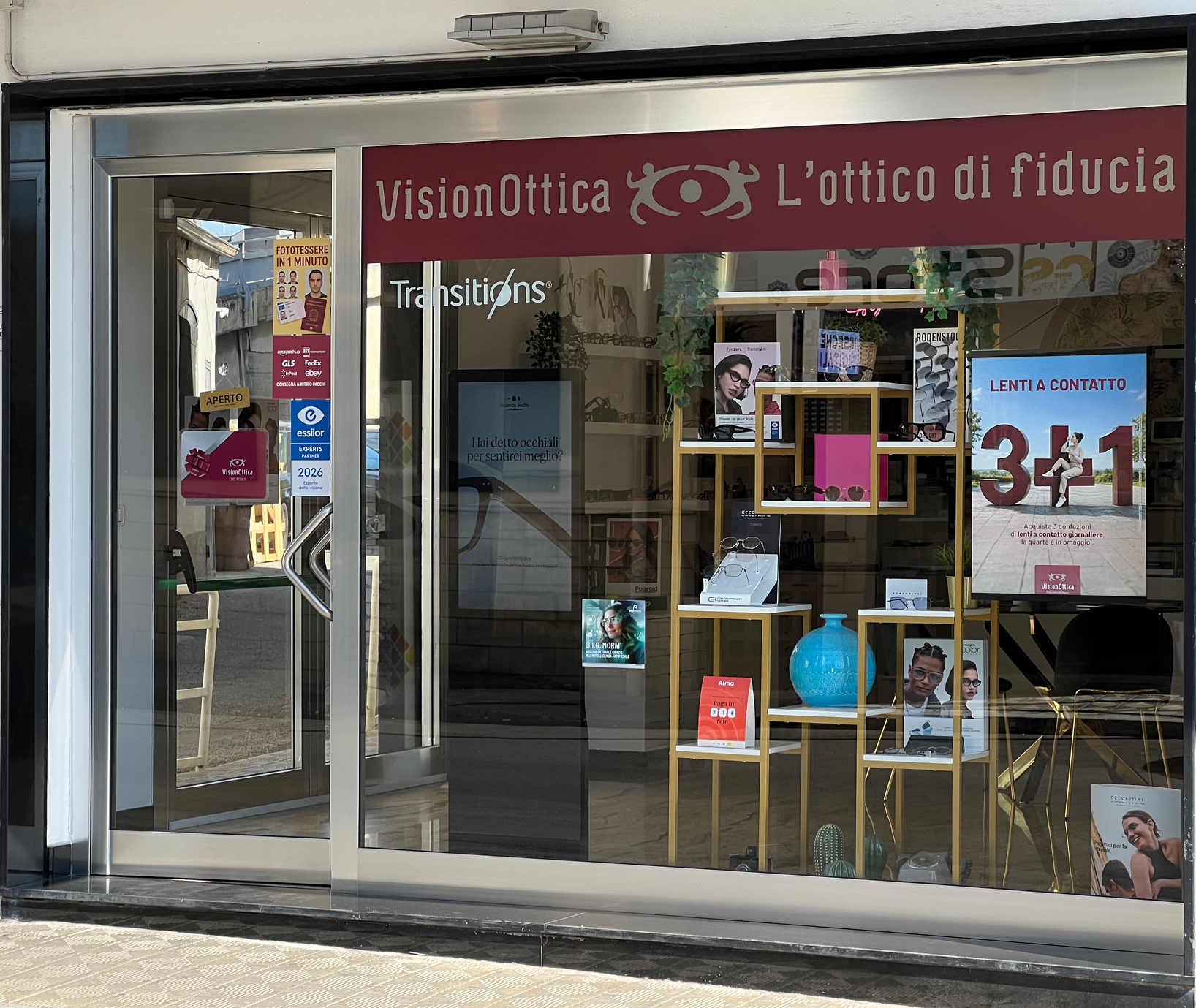 Vetrina del negozio VisionOttica L'Ottico di Fiducia di Reggio Calabria su strada con insegna bordeaux, porta automatica a vetri, espositore dorato con occhiali e banner Lenti a Contatto 3+1