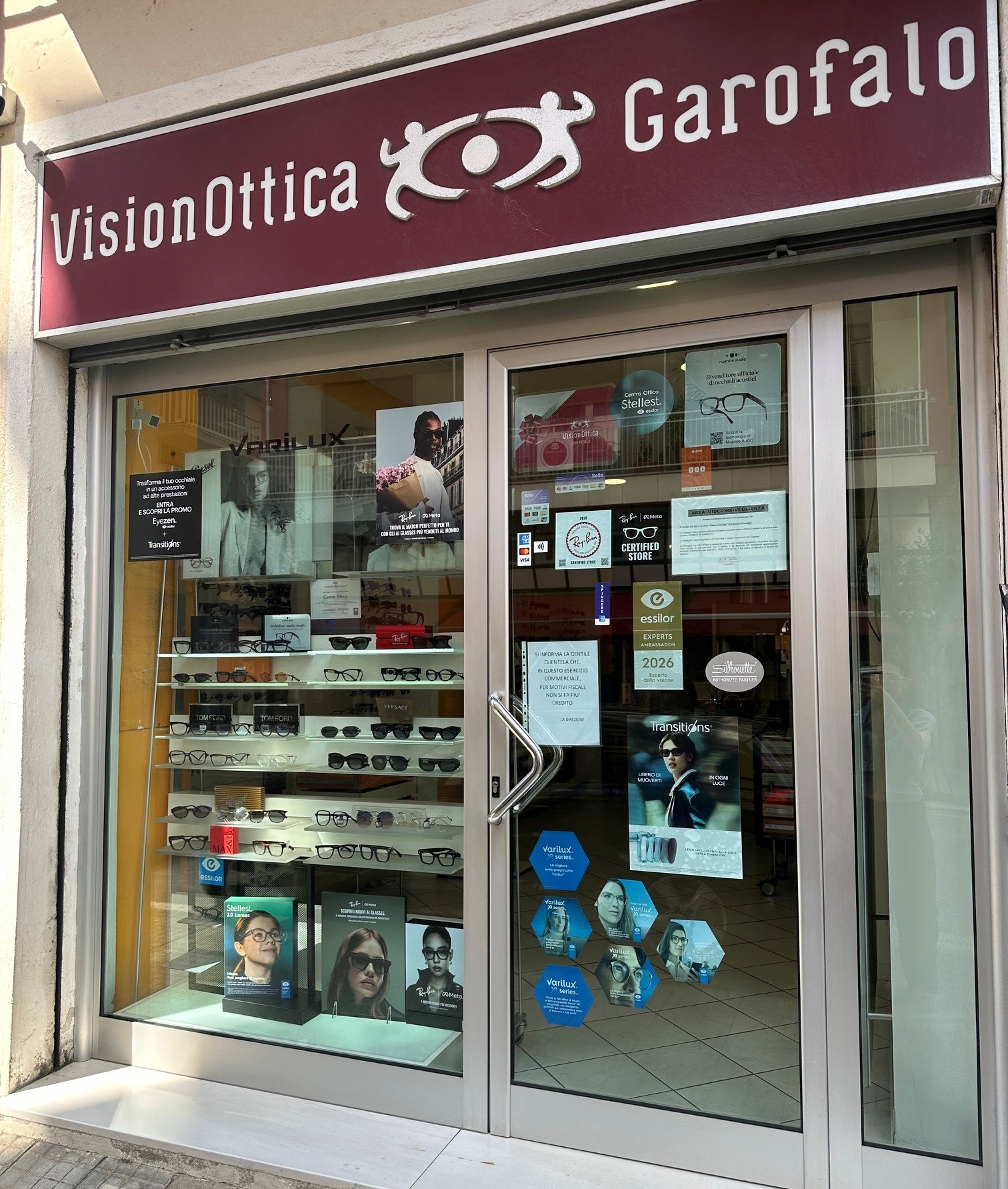 Vetrina del negozio VisionOttica Garofalo di Rogliano su strada con insegna bordeaux, porta a doppio battente con adesivi promozionali e scaffali di occhiali da sole e da vista