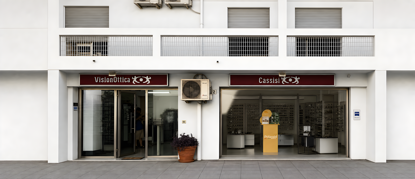 Facciata del negozio VisionOttica Cassisi di Cefalù su strada con edificio bianco, doppia insegna bordeaux, due aperture con porte scorrevoli a vetri e vaso di piante all'ingresso