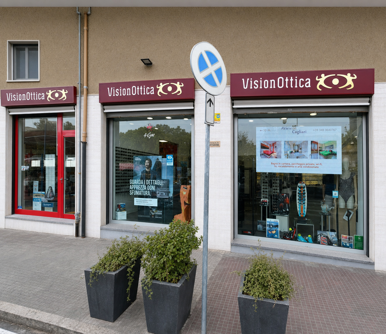 Facciata del negozio VisionOttica 4Eyes di Cagliari su strada con doppia insegna bordeaux, due vetrine con banner promozionali, porta rossa laterale e vasi di piante sul marciapiede