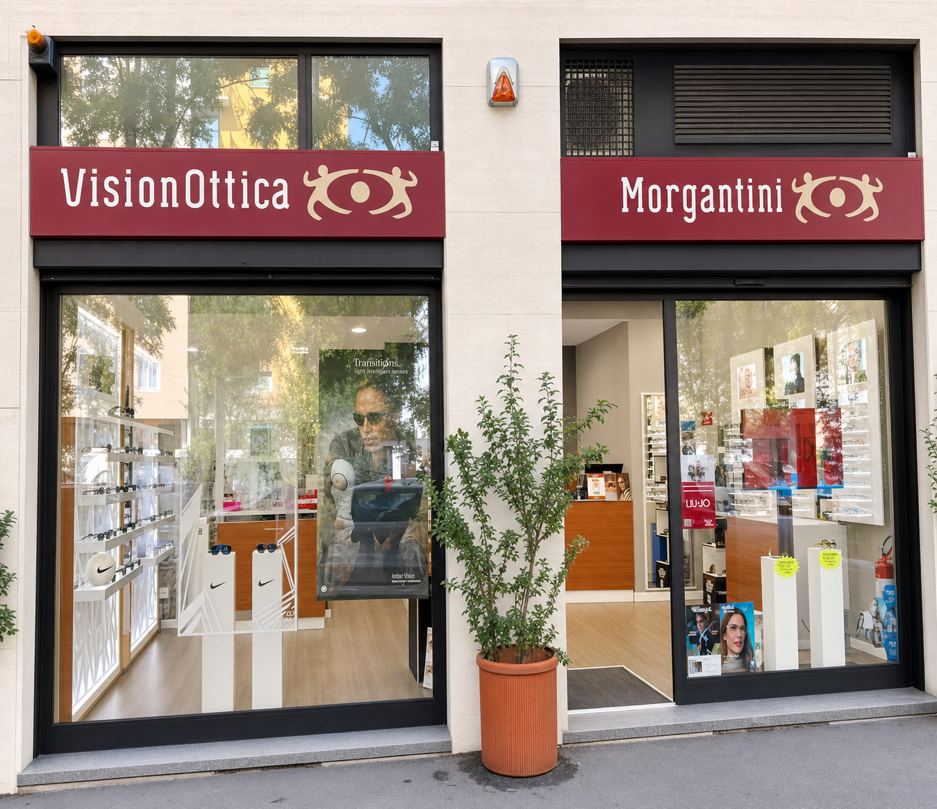 Facciata del negozio VisionOttica Morgantini di Milano su strada con doppia insegna bordeaux, vetrine ampie con espositori di montature e pianta decorativa all'ingresso