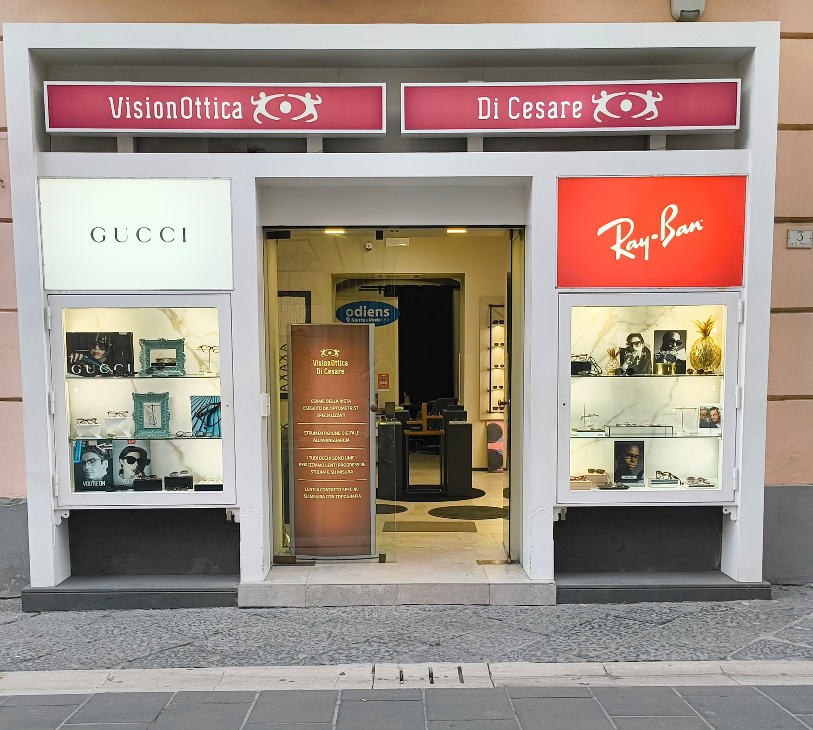 Facciata del negozio VisionOttica Di Cesare di Caserta su via pedonale con insegna bordeaux, vetrine laterali con logo Gucci e Ray-Ban e porta centrale a vetri aperta