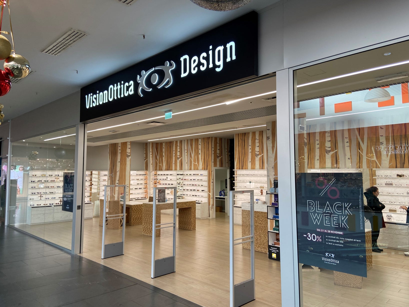 Ingresso del negozio VisionOttica Design di Asti in centro commerciale con insegna nera illuminata, interno a vista con espositori in legno chiaro e banner Black Week -30%