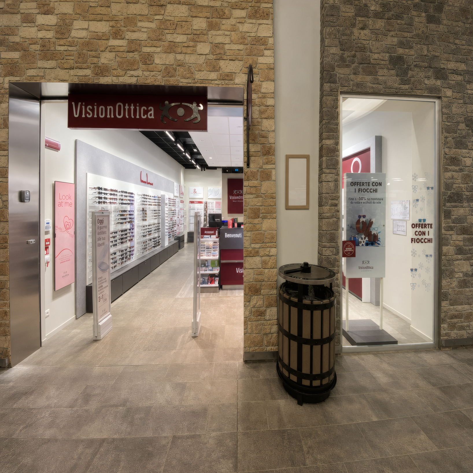 Ingresso del negozio VisionOttica Borgo d'Abruzzo di Cepagatti in centro commerciale con insegna bordeaux su pilastro, corridoio di passaggio con cestino e accesso laterale aperto