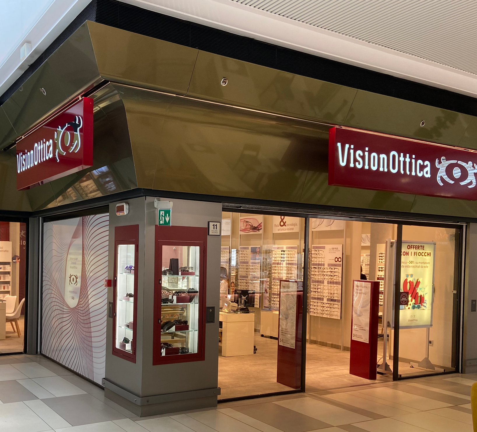 Ingresso del negozio VisionOttica Vialarga di Bologna in centro commerciale con doppia insegna bordeaux, pannello decorativo laterale, porta a vetri scorrevole e interno illuminato