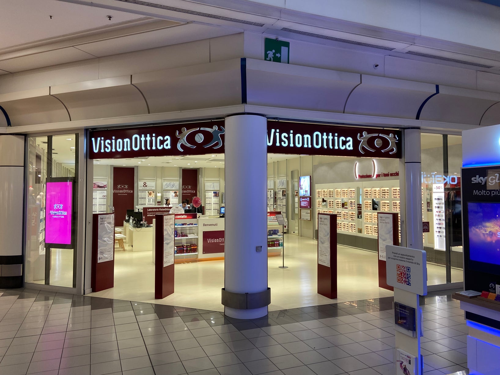 Ingresso del negozio VisionOttica Centrolame di Bologna in centro commerciale con doppia insegna retroilluminata bordeaux, ampio varco aperto, espositori laterali e schermo digitale all'interno