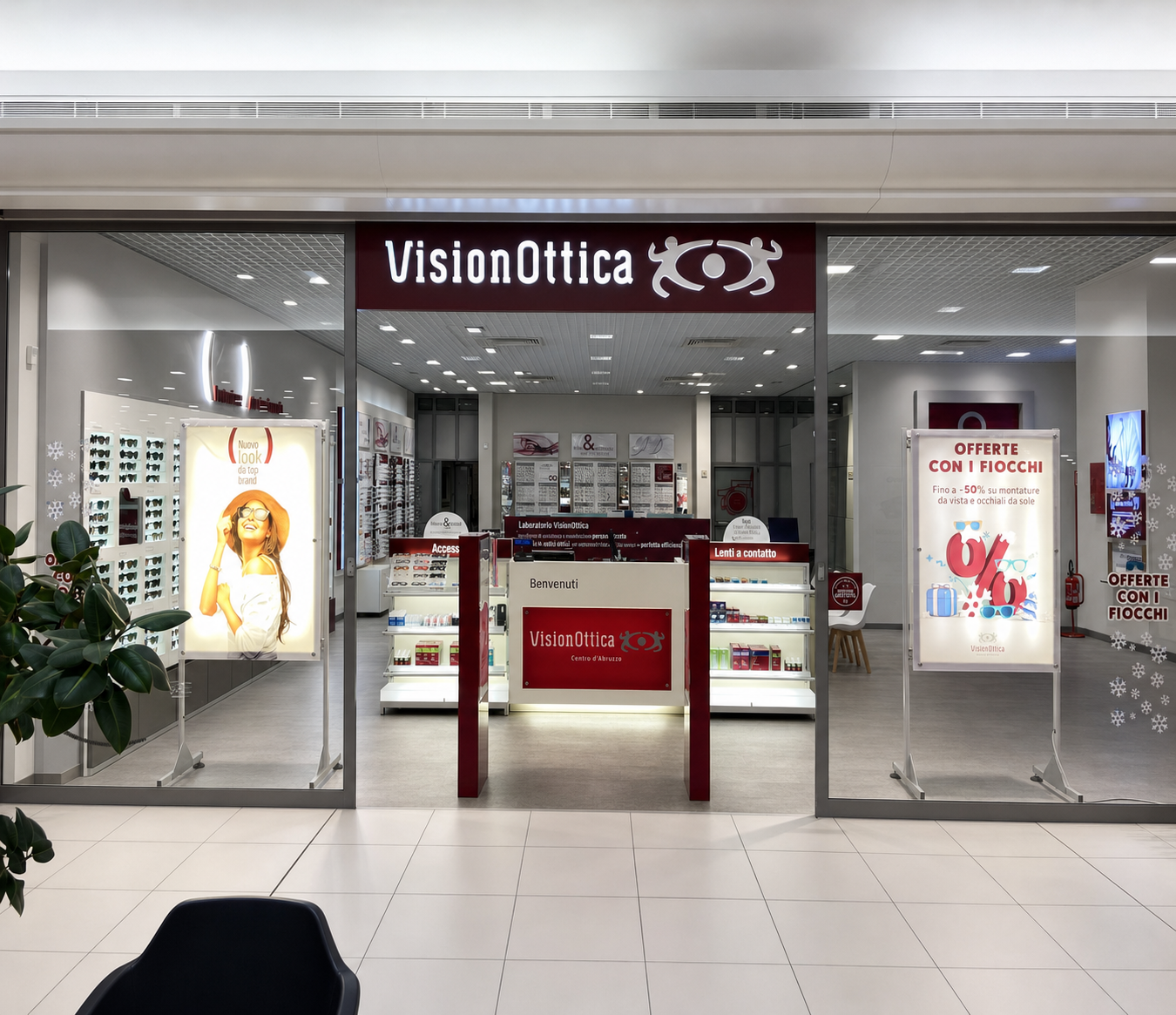 Ingresso del negozio VisionOttica Centro d'Abruzzo di San Giovanni Teatino in centro commerciale con insegna bordeaux, vetrine laterali con poster e totem promozionali e bancone interno visibile